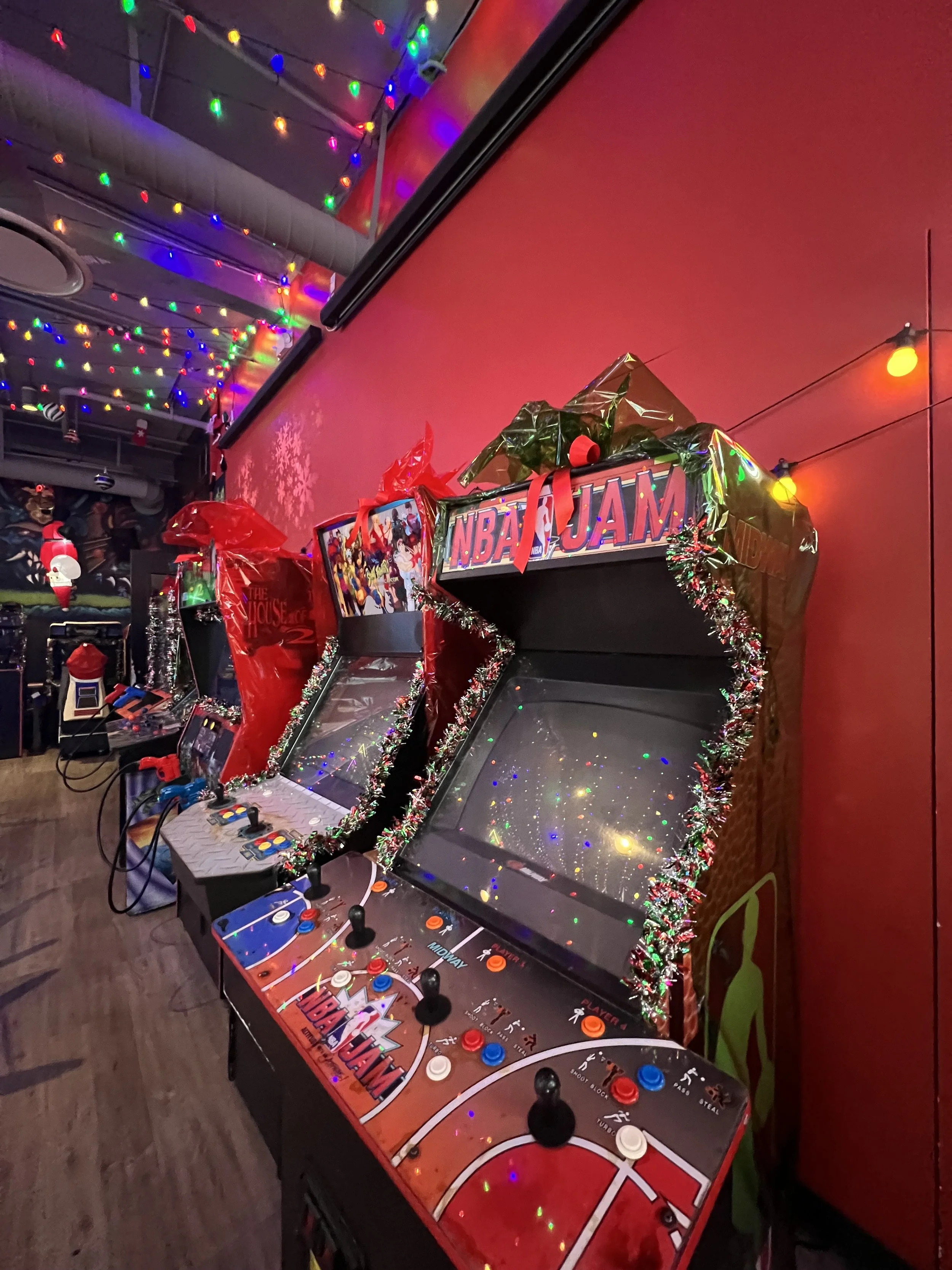 Three vintage arcade basketball games decorated with holiday tinsel and Christmas-themed embellishments, with colorful string lights hanging from the ceiling. Mini Boss San Jose