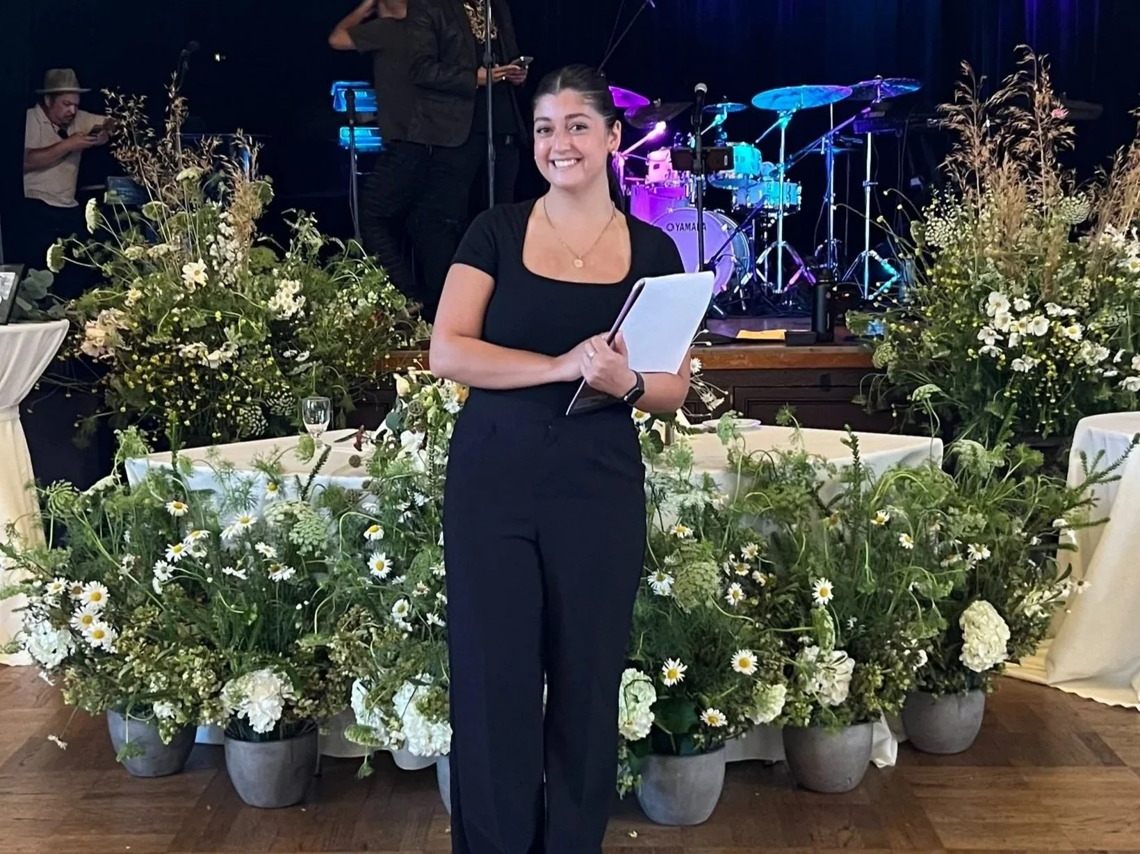 A woman in black pants and top stands smiling in front of a floral arrangement holding a notepad. In the background, there is a stage with musical instruments and two people standing, one using a phone.