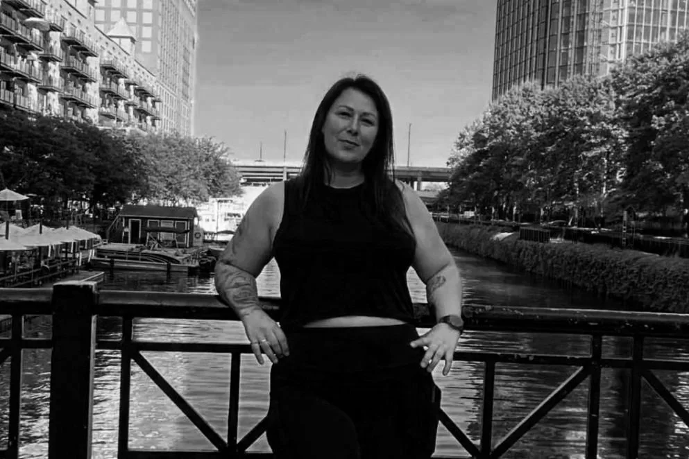 A woman with dark hair and tattoos standing by a canal in an urban area with high-rise buildings and trees.