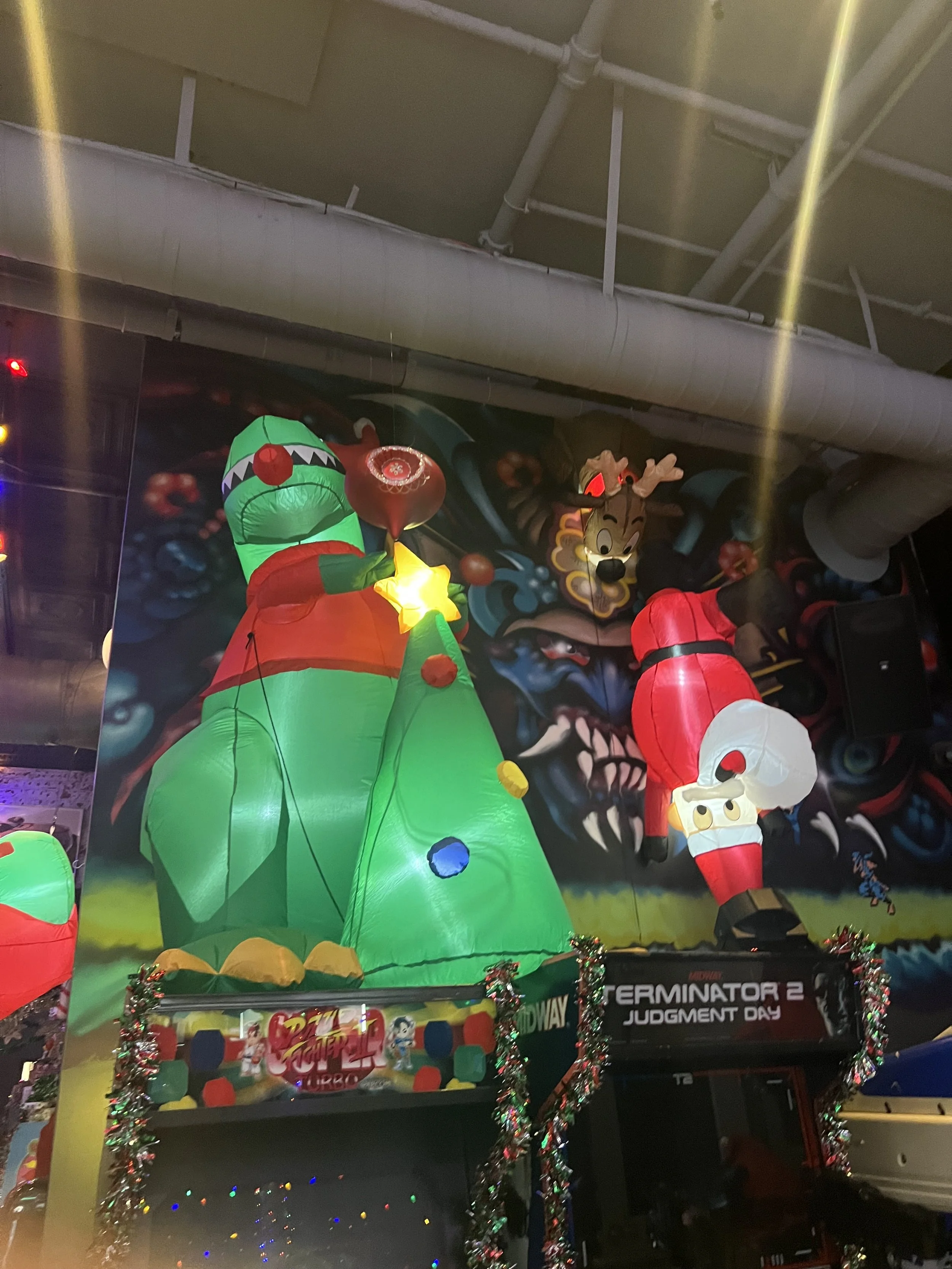 Colorful holiday decorations featuring a large inflatable Christmas tree with a star on top, a frog figure with a red hat, and a Santa figure, with a mural of a fierce creature in the background, and a sign that reads 'Terminator 2 Judgment Day' deco