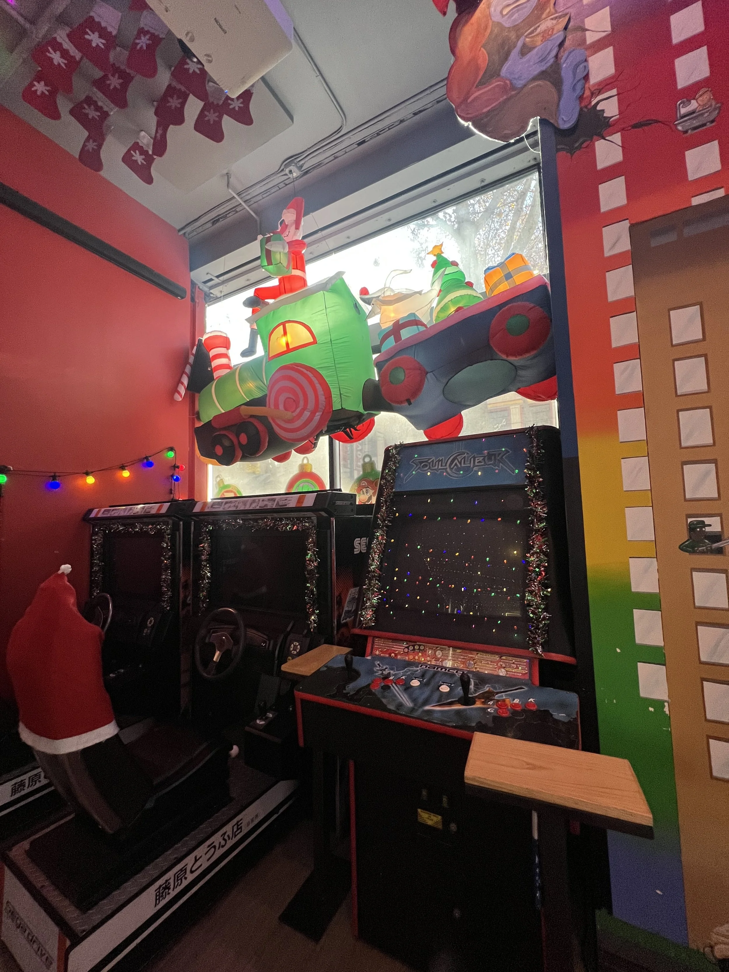 Indoor arcade with Christmas-themed decorations, including inflatable decorations on the window, arcade game machines, and a string of colorful holiday lights. Mini Boss San Jose. 