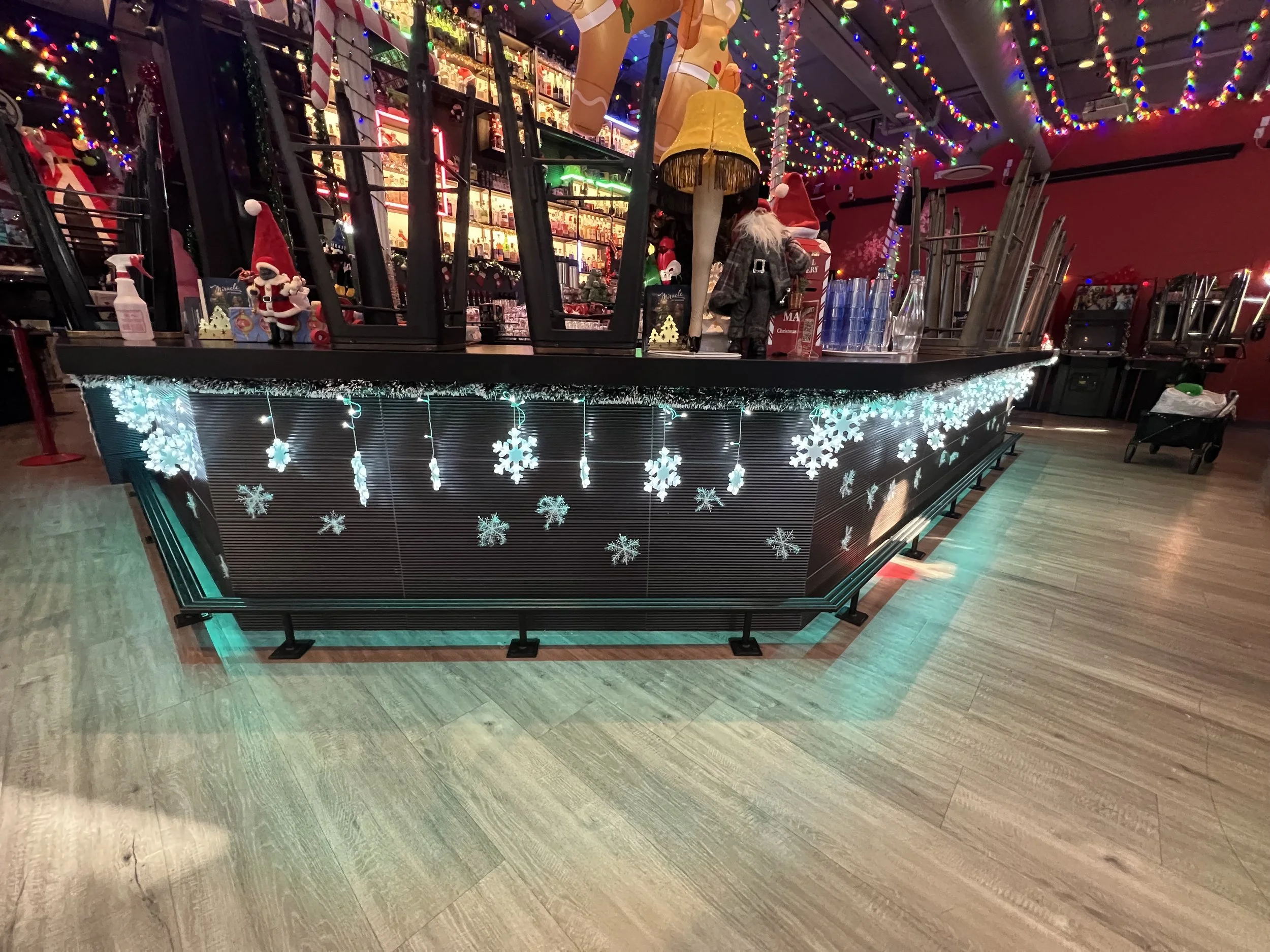 A decorated bar or counter area with Christmas decorations, including snowflake string lights and ornaments. The area is topped with stacked chairs and some holiday figures, all illuminated by colorful string lights overhead. Mini Boss San Jose.
