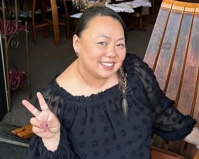 A woman smiling and making a peace sign with her fingers, sitting in a restaurant or café.