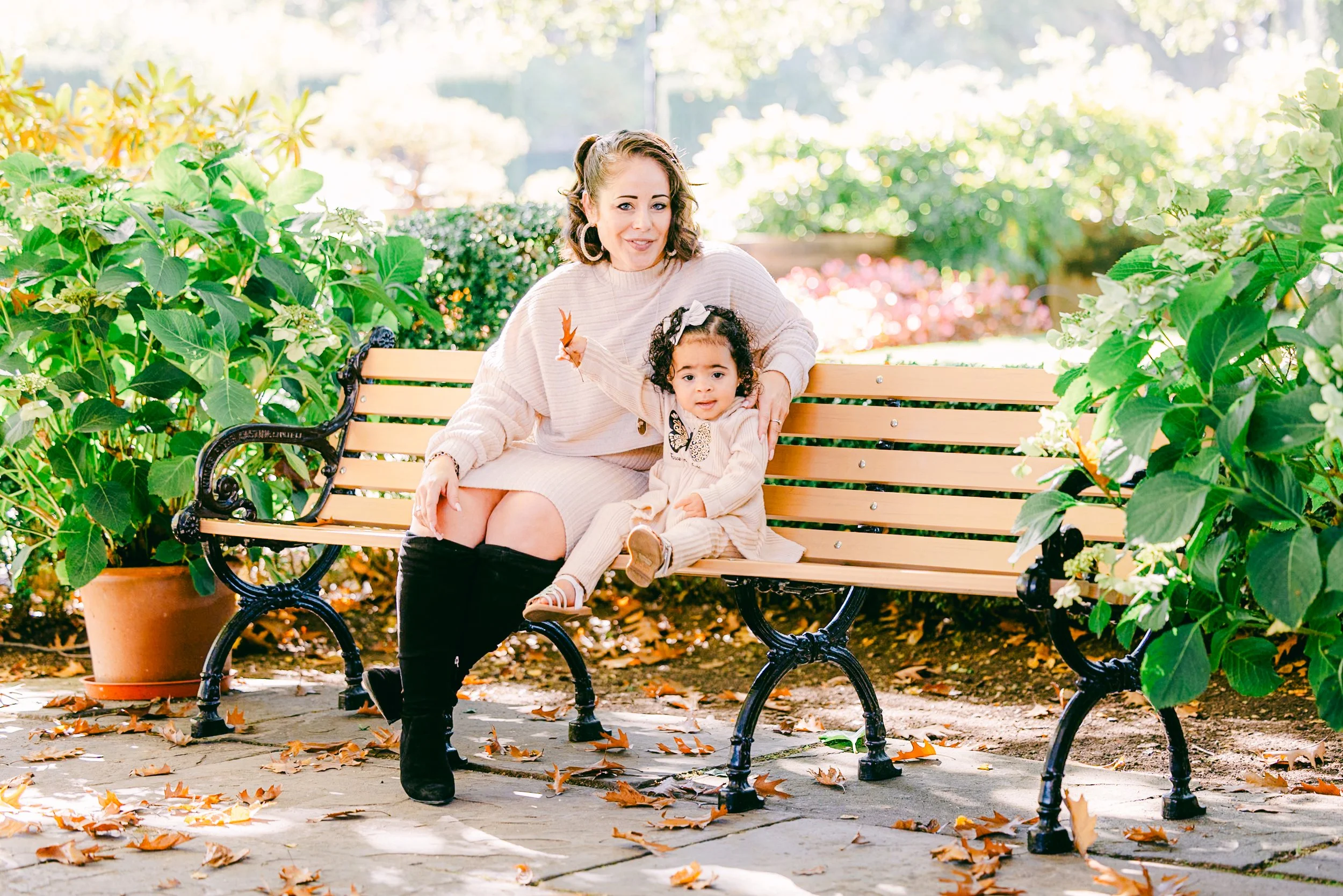 A woman and a young girl sitting on a park bench surrounded by lush greenery and plants, with fallen autumn leaves on the ground, outdoors on a sunny day.