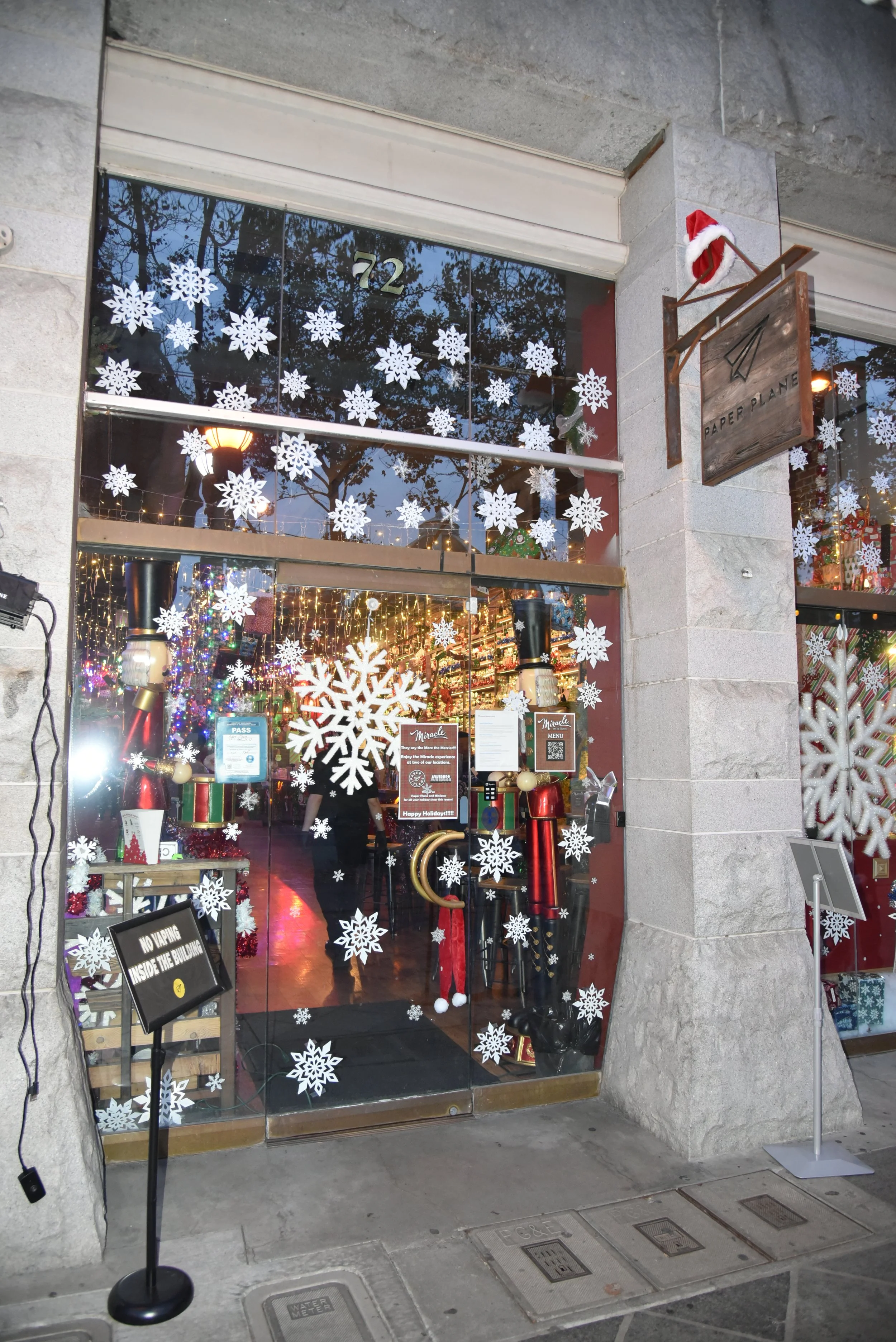 Storefront decorated with Christmas snowflake window decals, a Santa hat on a hanging sign, and Christmas ornaments inside. Paper Plane San Jose.