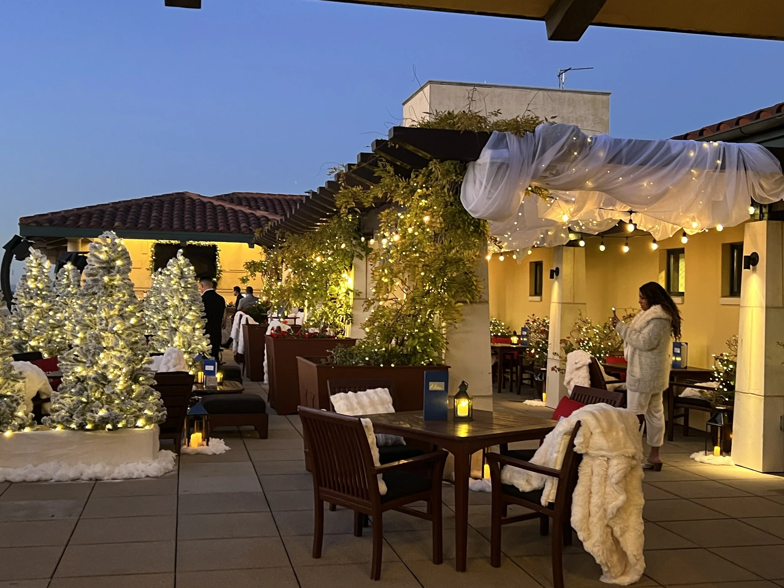 A decorated outdoor rooftop patio at dusk with white Christmas trees, string lights, candles, and people in warm clothing, as well as tables and chairs.