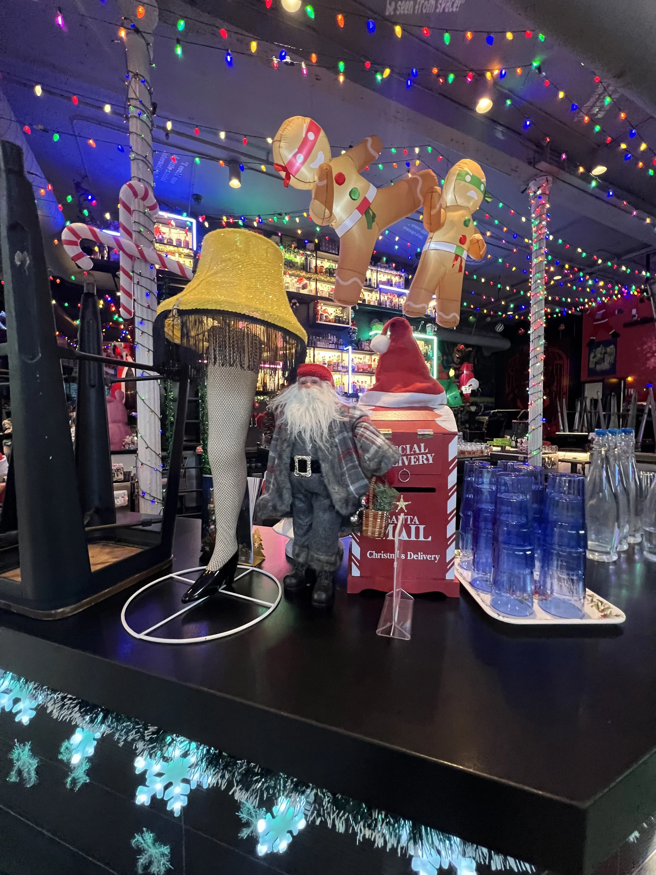 Decorative Christmas scene with inflatable gingerbread men, Santa figurine, and holiday lights, set on a bar counter with glasses and a tray. Mini Boss San Jose.