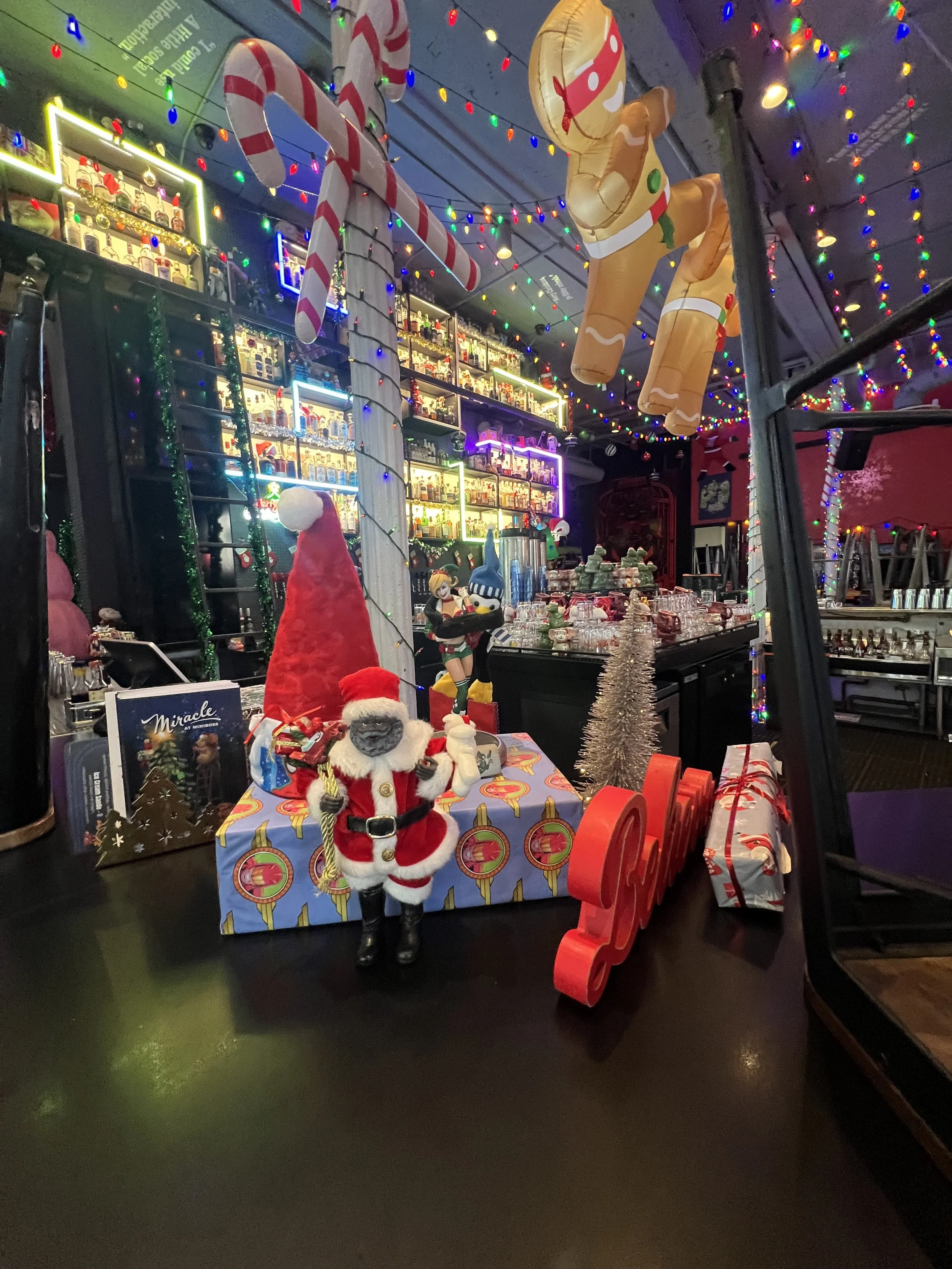 Festive Christmas decorations with lights, a small Santa figure on a gift-wrapped box, a red sleigh, holiday ornaments, and hanging inflatable gingerbread and candy cane decorations inside a decorated bar or restaurant. Mini Boss San Jose. 