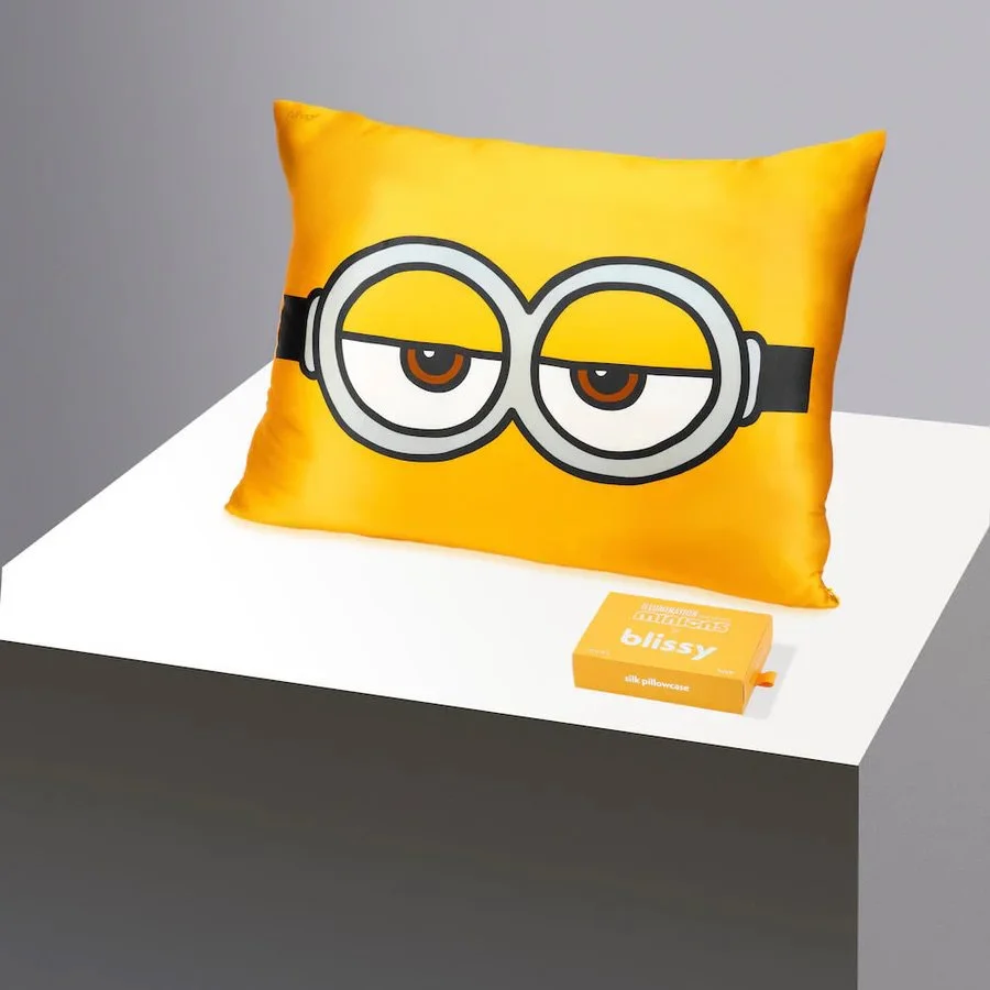 A rectangular, yellow pillow with a graphic of a Minions character's face, showing half-lidded eyes and the goggles in black and white. The pillow is placed on a white surface with a gray background. A small yellow box labeled 'blissy' is next to it.
