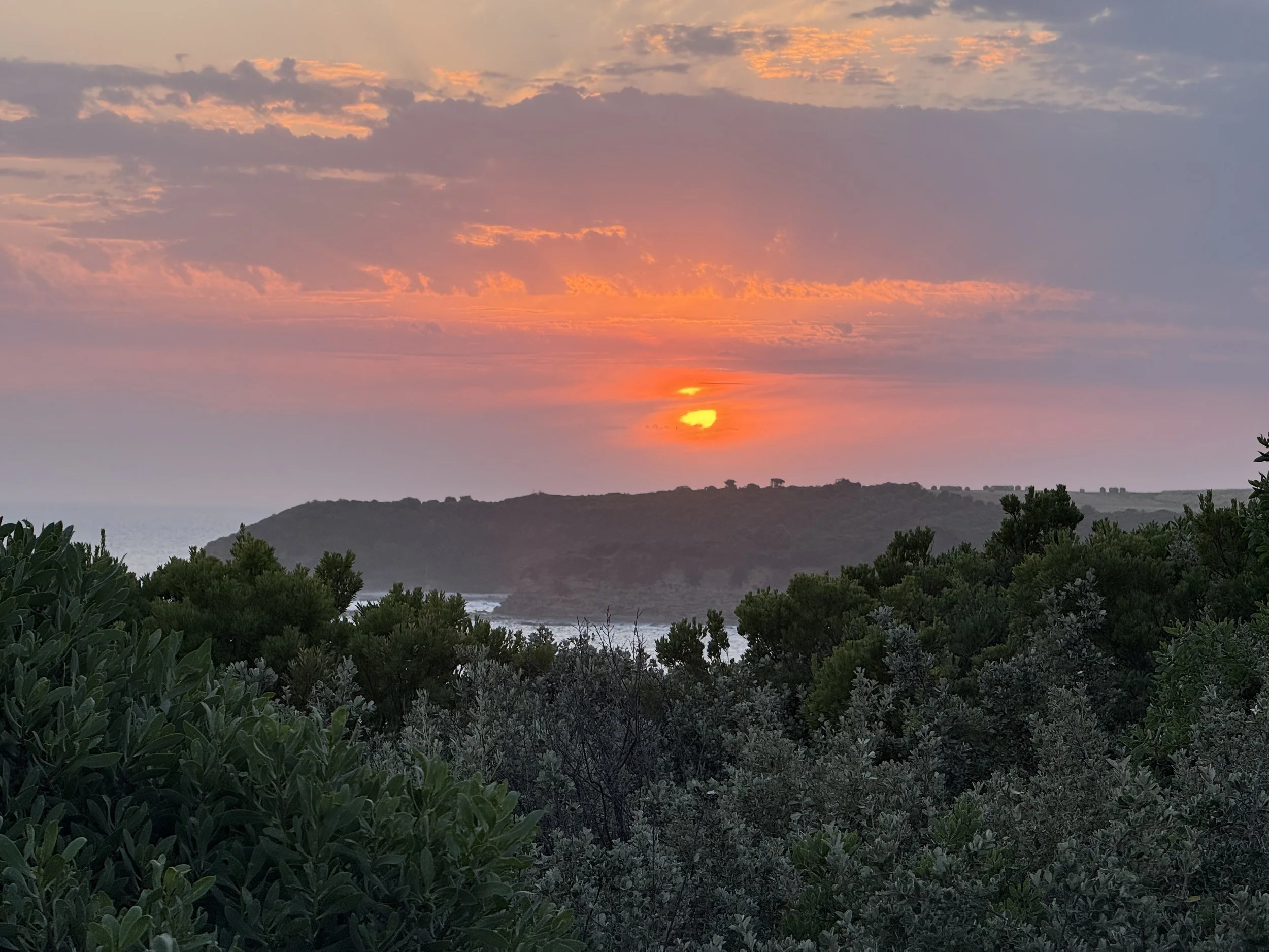 Sunset over a hillside with clouds in the sky, overlooking a body of water and green foliage in the foreground.