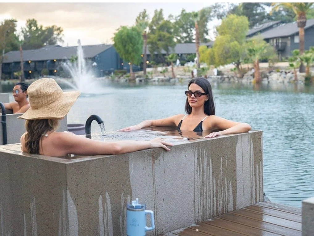 Two women relaxing in a concrete hot tub on a dock by a lake, with trees and buildings in the background. One woman is wearing a large sun hat, the other is wearing sunglasses.