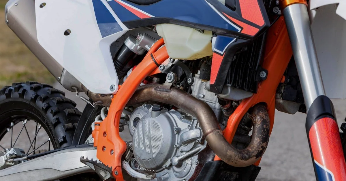 A closeup of a dirt bike with the metal engine exposed beneath a red, blue, and gray body. It is sitting outside.