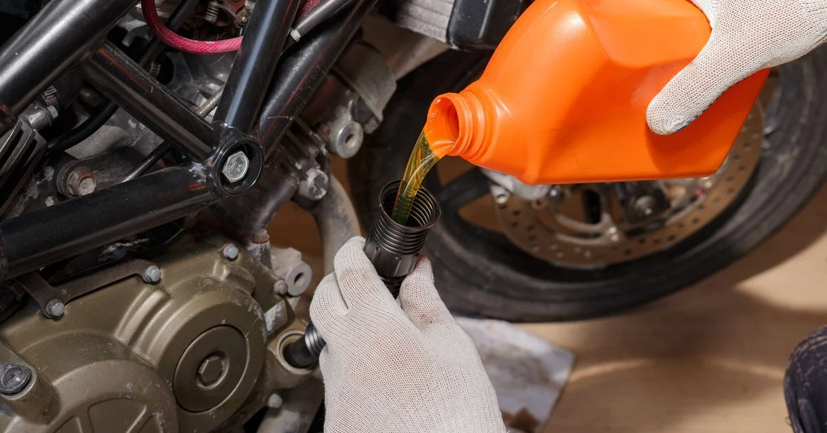 A person wearing white gloves is pouring an orange bottle of oil into the engine of a motorcycle.