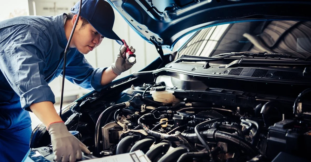A mechanic wearing a blue hat and coveralls holding a flashlight while inspecting the engine bay of a car.