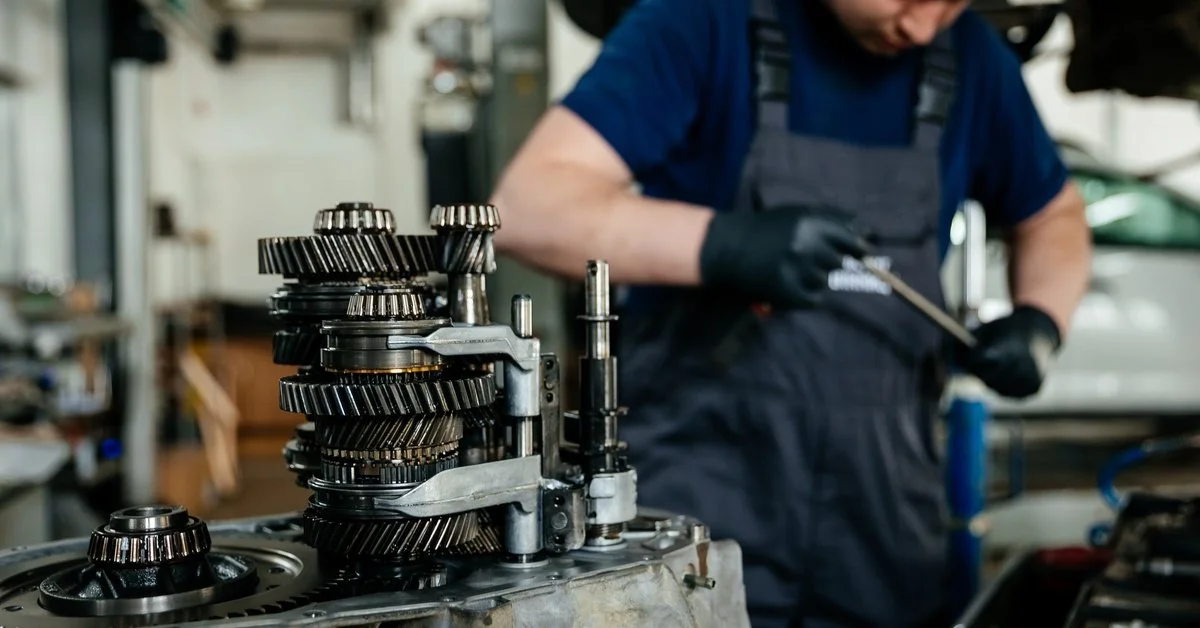 A manual gearbox in focus in the foreground with a mechanic using a tool in a shop in the background.