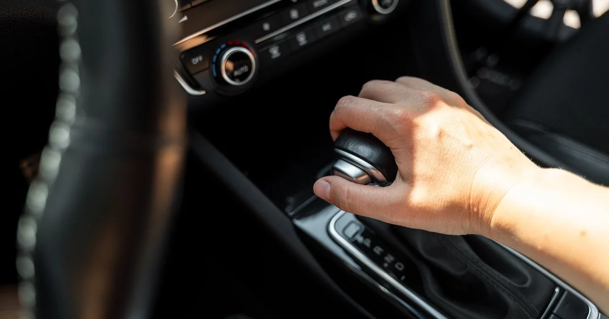A close-up of a person holding onto the transmission shifter in a car while sitting in the driver’s seat.