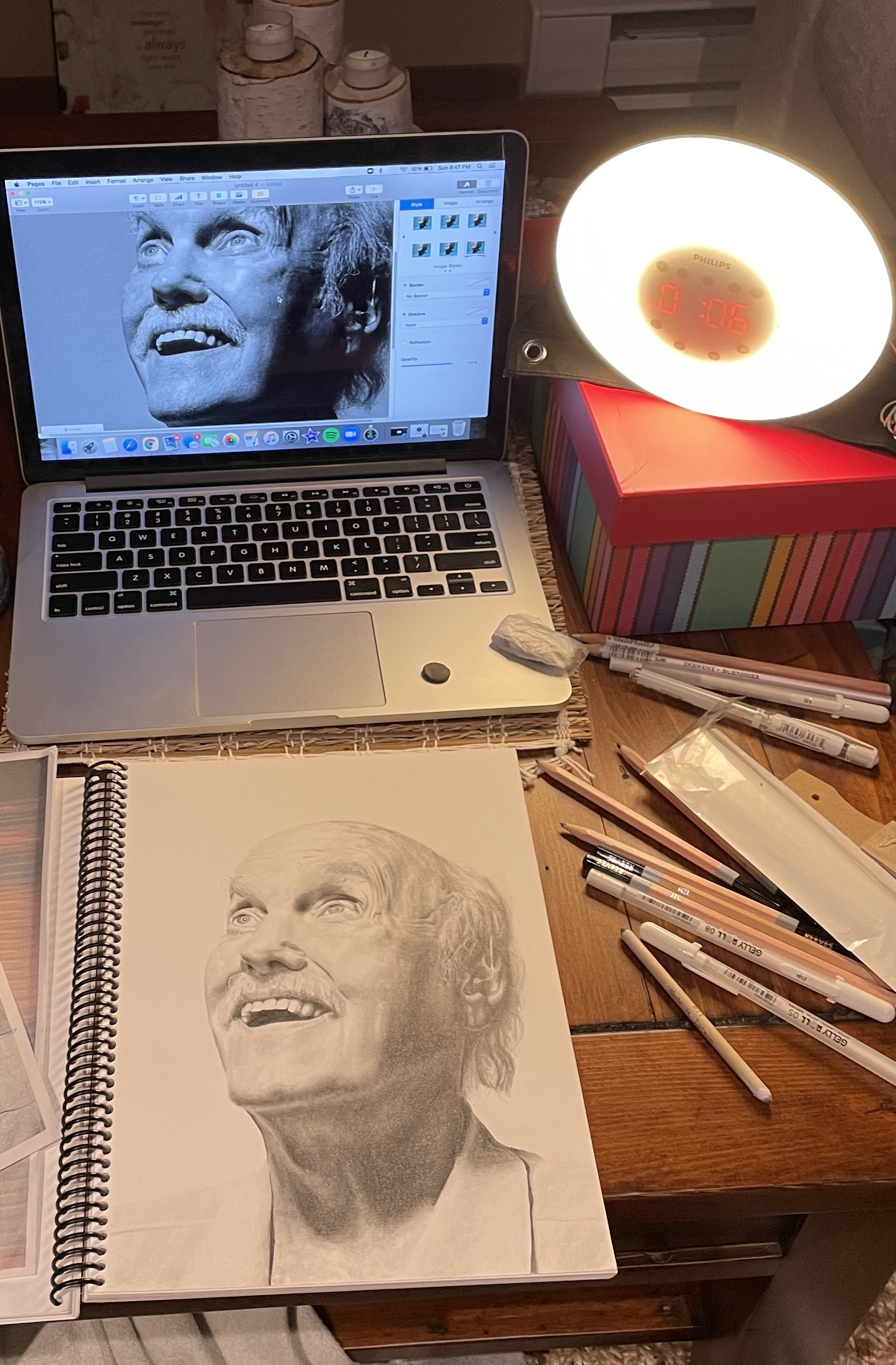 A photo of the artist's desk/workspace. There is a laptop, a bright lamp, and a sketchbook with a few pens scattered next to it.