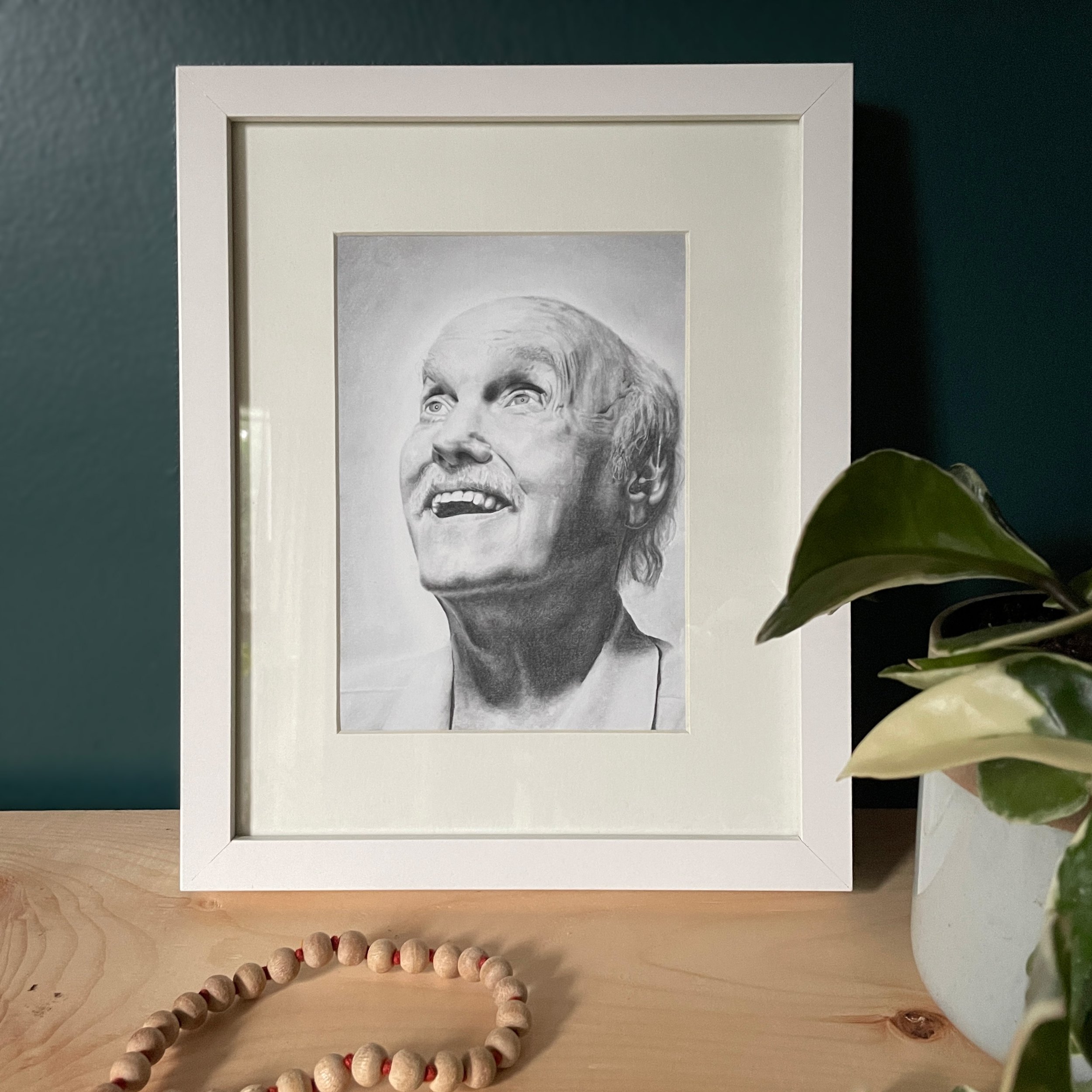 A photo of a framed art print of Ram Dass—it is sitting on a pine wood shelf alongside a houseplant and a mala.
