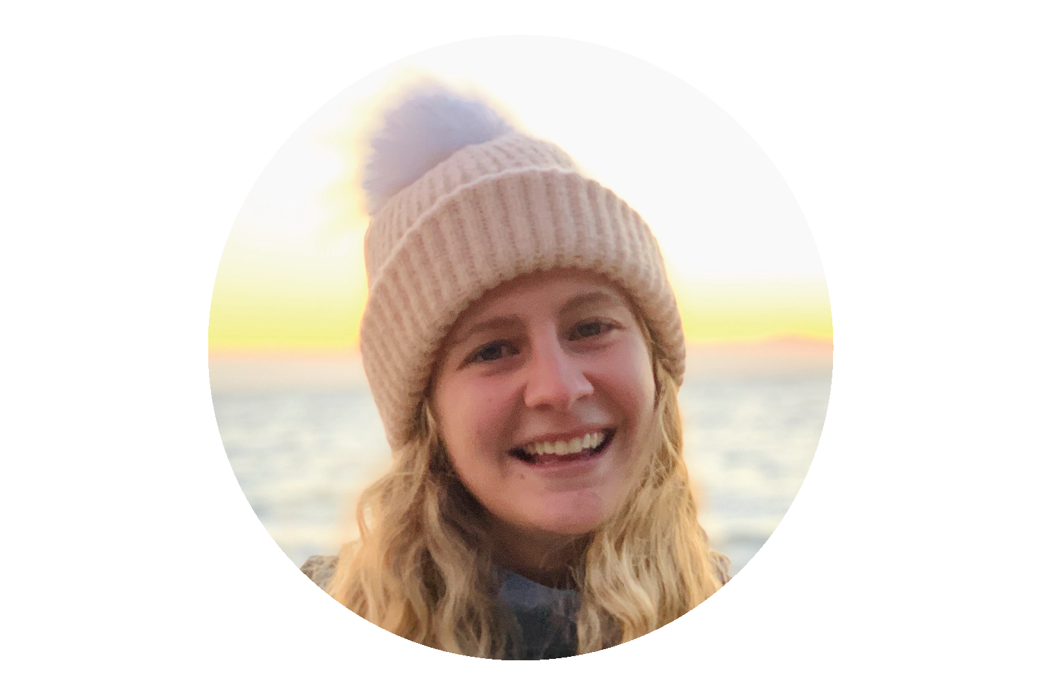 A photo of a smiling Lakshmi with the ocean in the background. She is wearing a light pink knit hat with a big fuzzy pom pom.