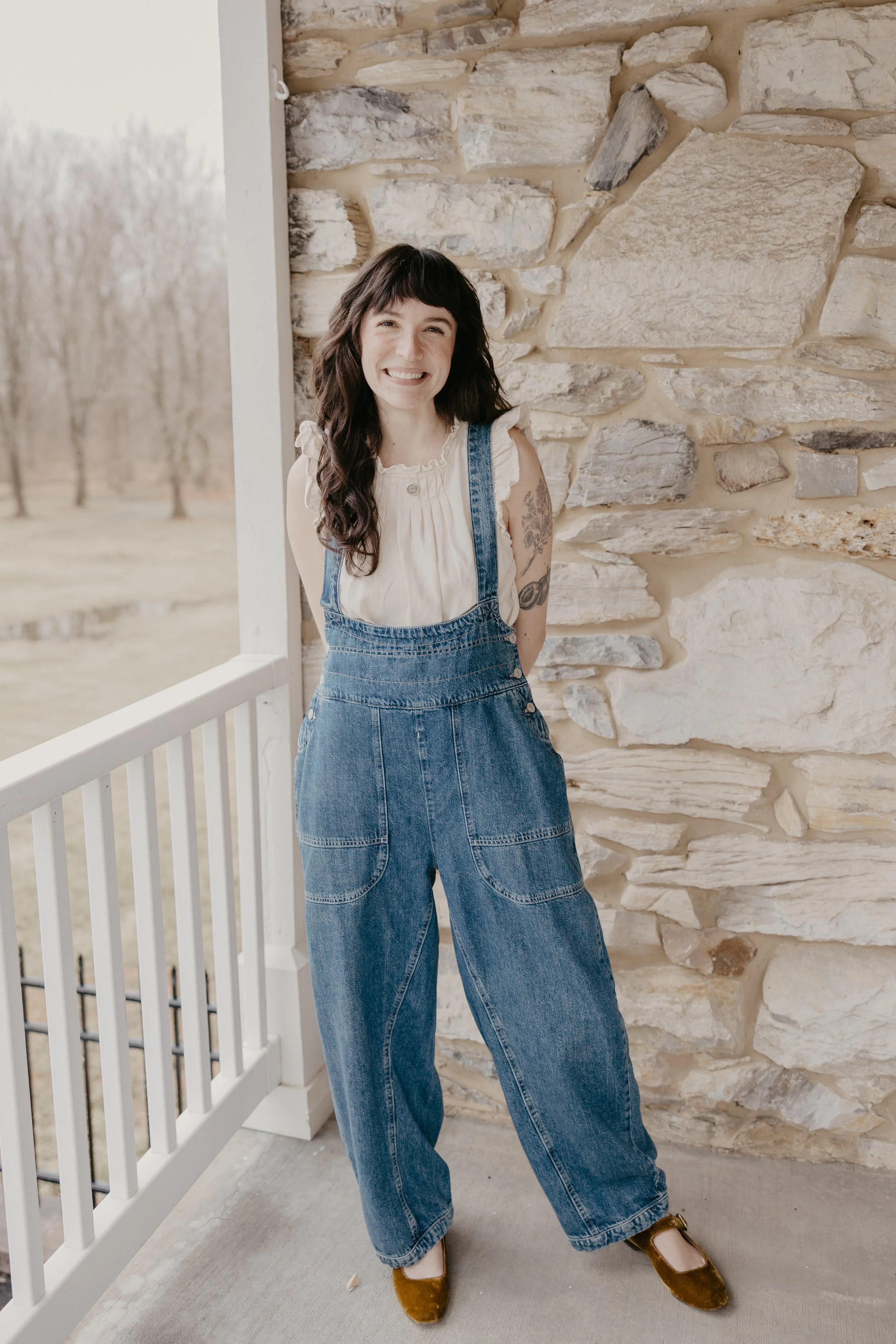 Madison, an art therapist, with dark hair and tattoos, smiling, standing on the porch of The Art Room, with a stone wall behind her, wearing overalls, a cream blouse, and brown velvet shoes.