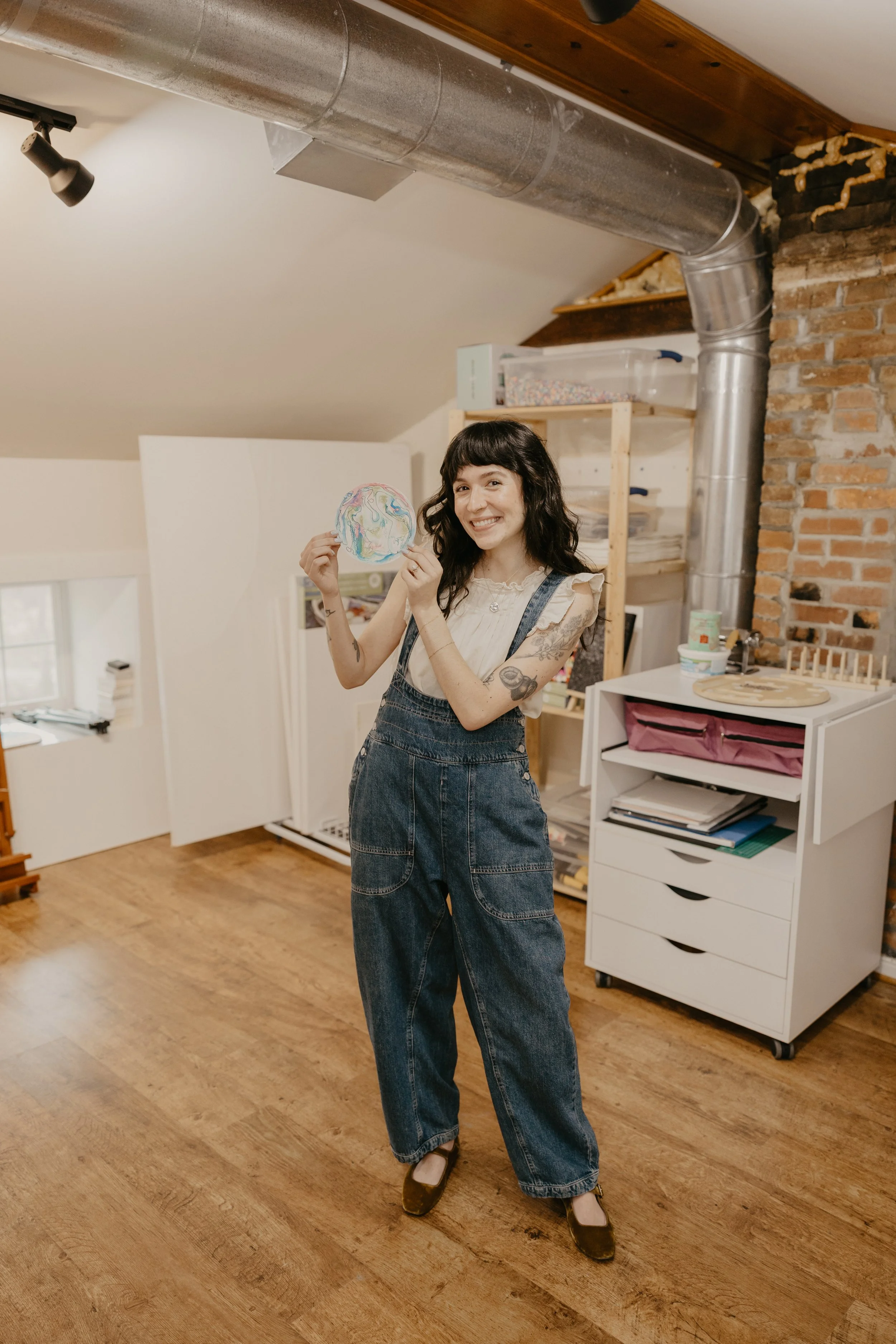 Madison, an art therapist with dark hair and tattoos, wearing overalls, is smiling and holding a colorful, circle mandala artwork in the creative workspace of our office, The Art Room, with wooden flooring, brick wall, and storage shelves.