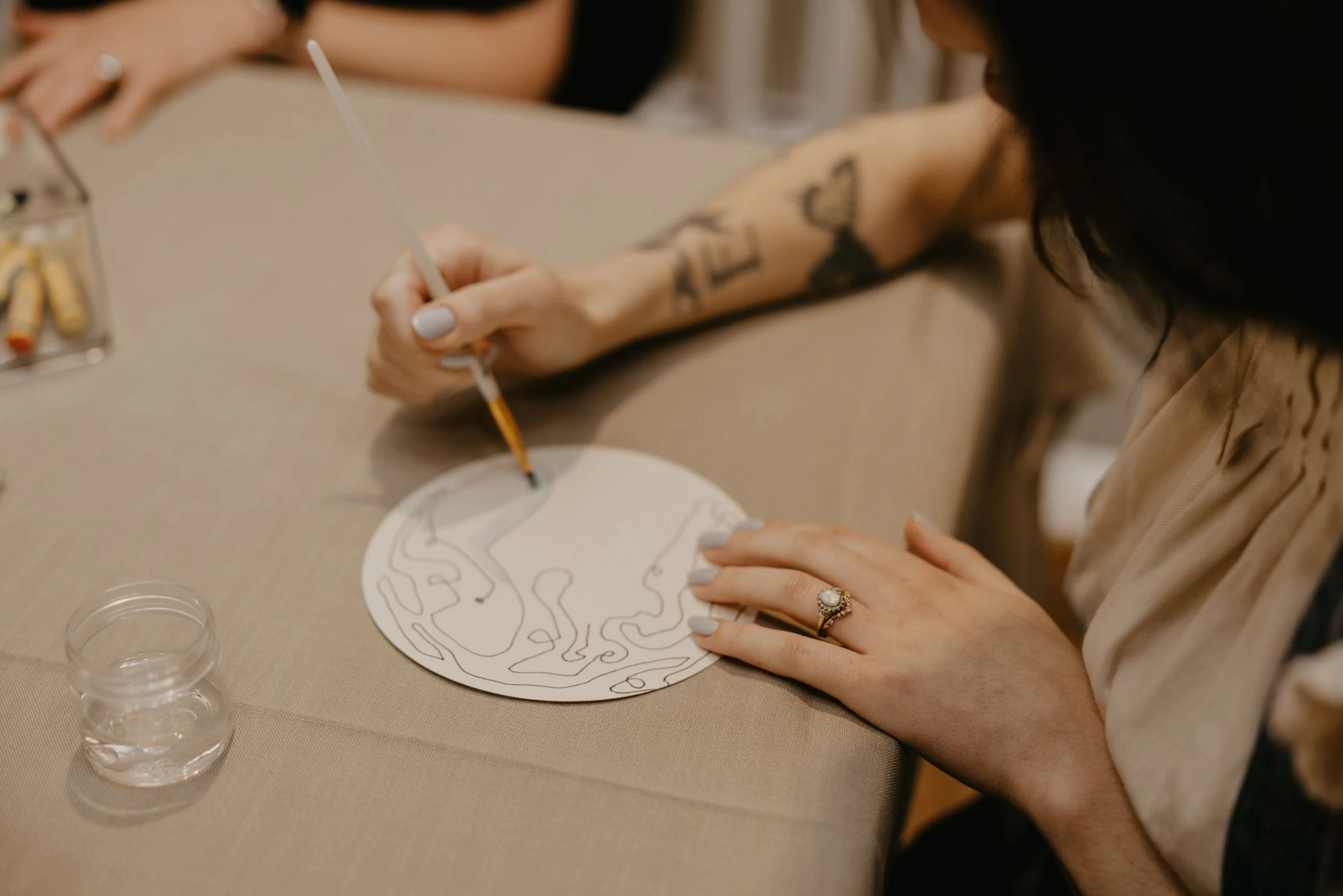 Madison's, an art therapist, arm with tattoos, painting on a circular piece of watercolor paper with a small paintbrush, sitting at a beige table with a small glass of water nearby.