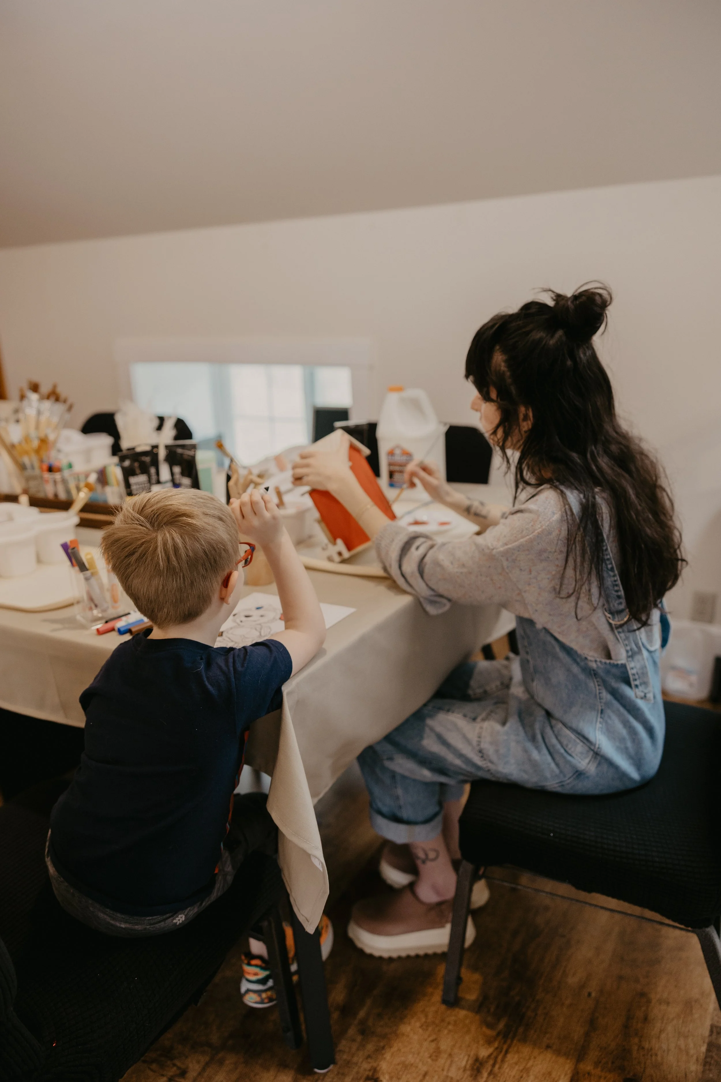 Madison, an art therapist, and a boy sitting at a table, engaging in arts and crafts activities with markers and paper, in the group space of The Art Room, a cozy room with a sloped ceiling and a window in the background.