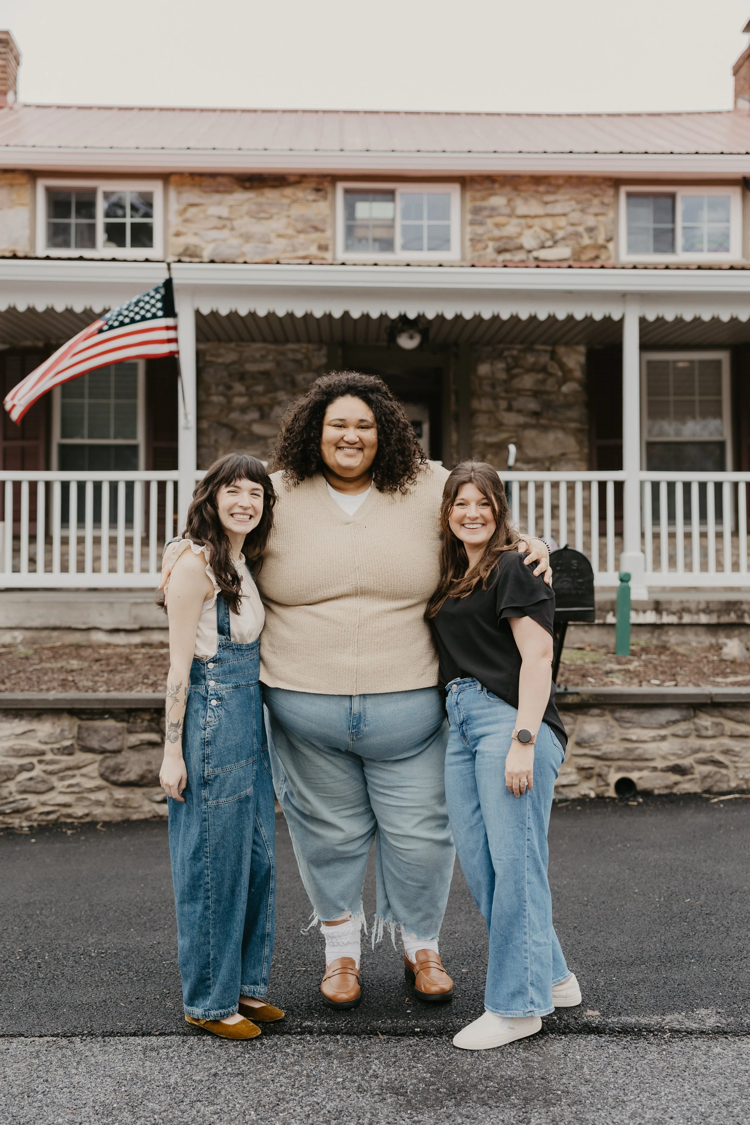 Three women art therapists, Madison, Claire, and Lauren, standing in front of The Art Room, a stone building with a porch, smiling and embracing each other.