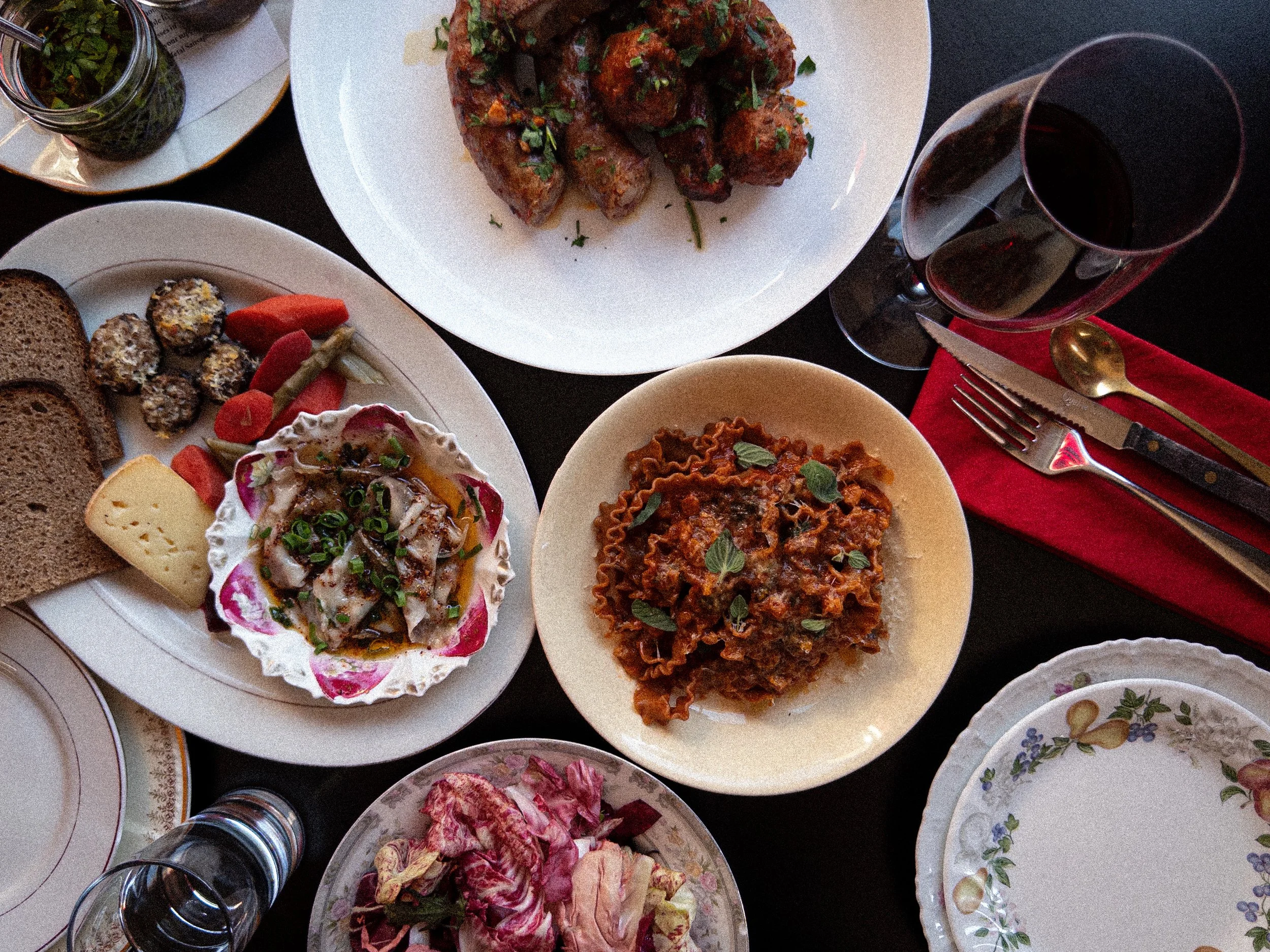 A variety of dishes on a table, including a plate of spaghetti with meat sauce, a bowl of pasta, a salad with vegetables and dressing, slices of bread, and glasses of red wine.