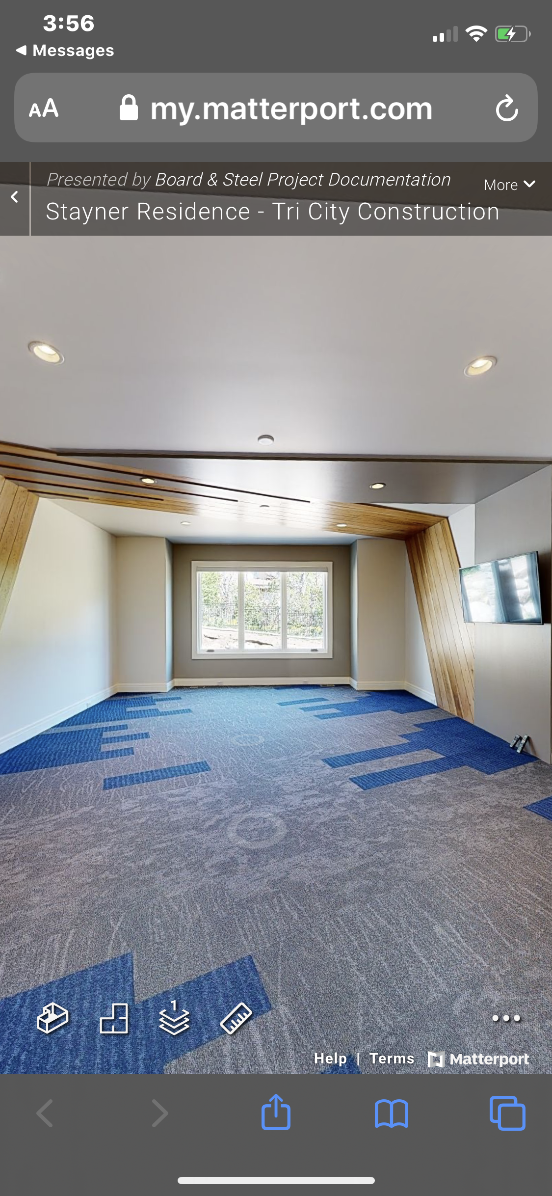 An empty room with beige walls, a large window, blue patterned carpet, and wooden accents on the ceiling and one wall, with a wall-mounted TV on the right.