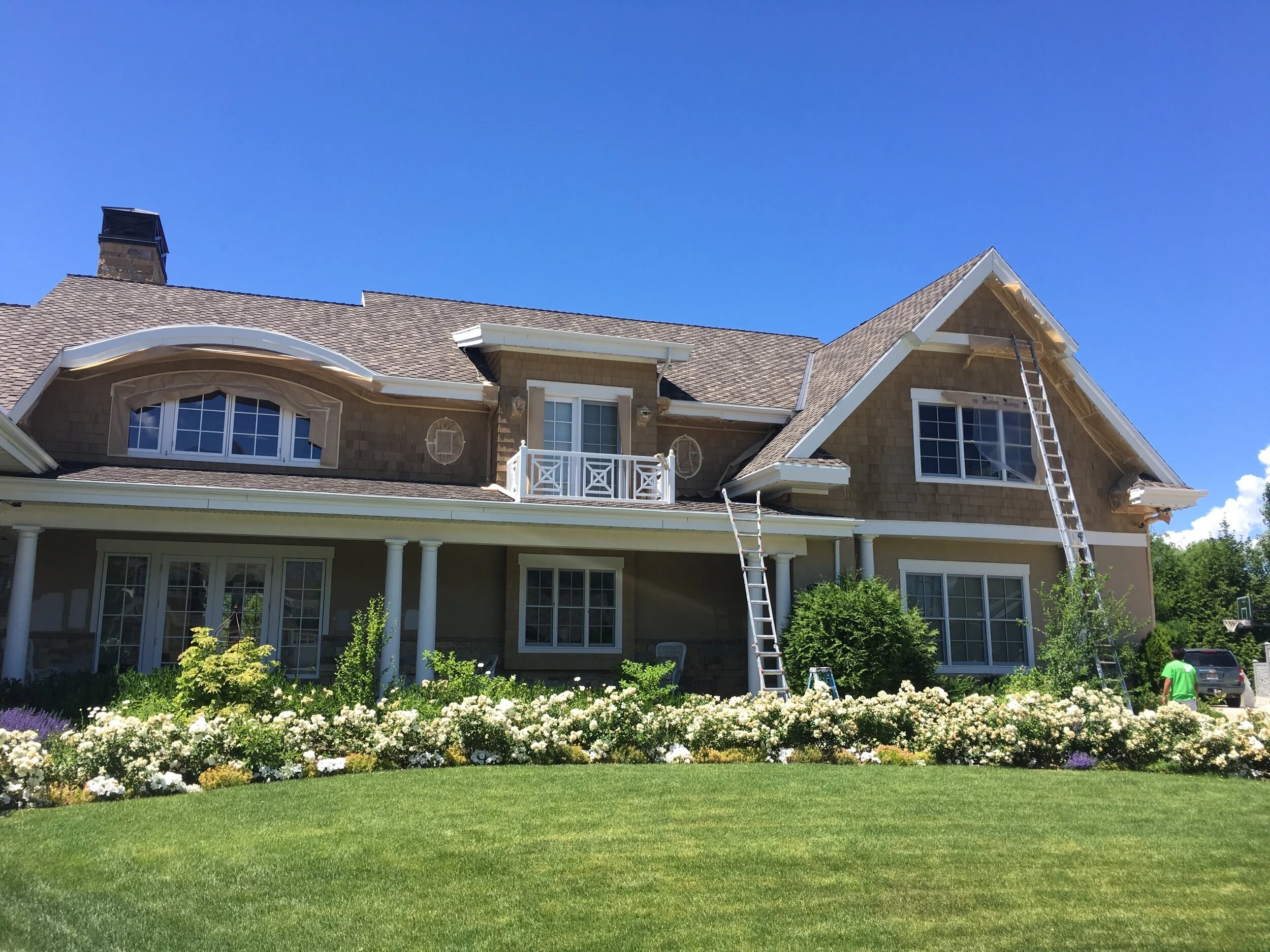 A large house under construction with ladders, surrounded by a lush green lawn and garden with blooming flowers and shrubs, under a clear blue sky.