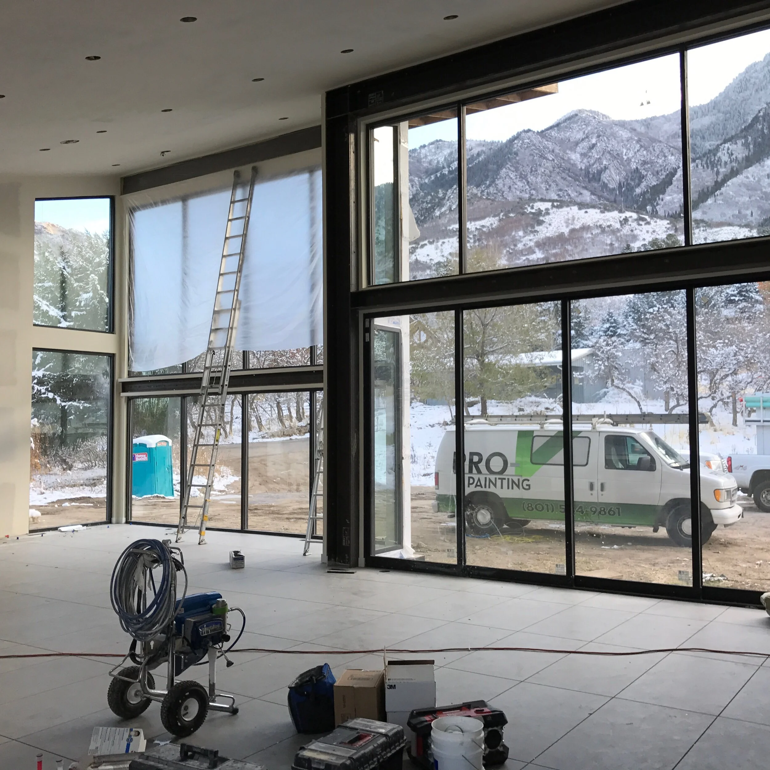 Interior construction site with large floor-to-ceiling windows showing snow-covered mountains outside. There is a ladder, construction tools, and materials scattered on the floor, indicating ongoing work.