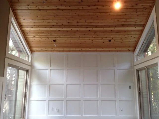 Interior of a room under construction with a wooden ceiling, white paneled walls, multiple electrical outlets, and large windows on two sides.