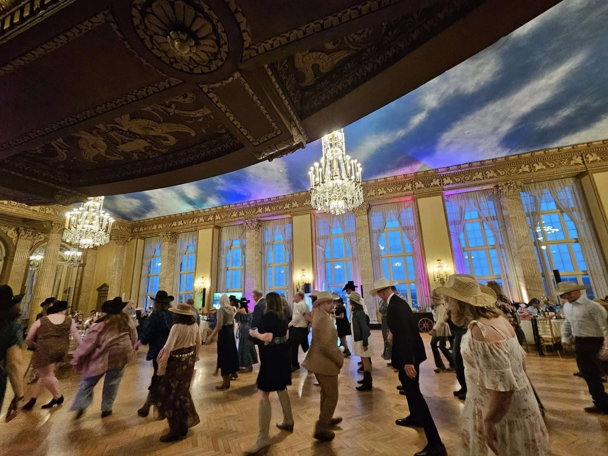 People dancing in a large ballroom with ornate decorations, chandeliers, tall windows, and a painted ceiling depicting a sky.