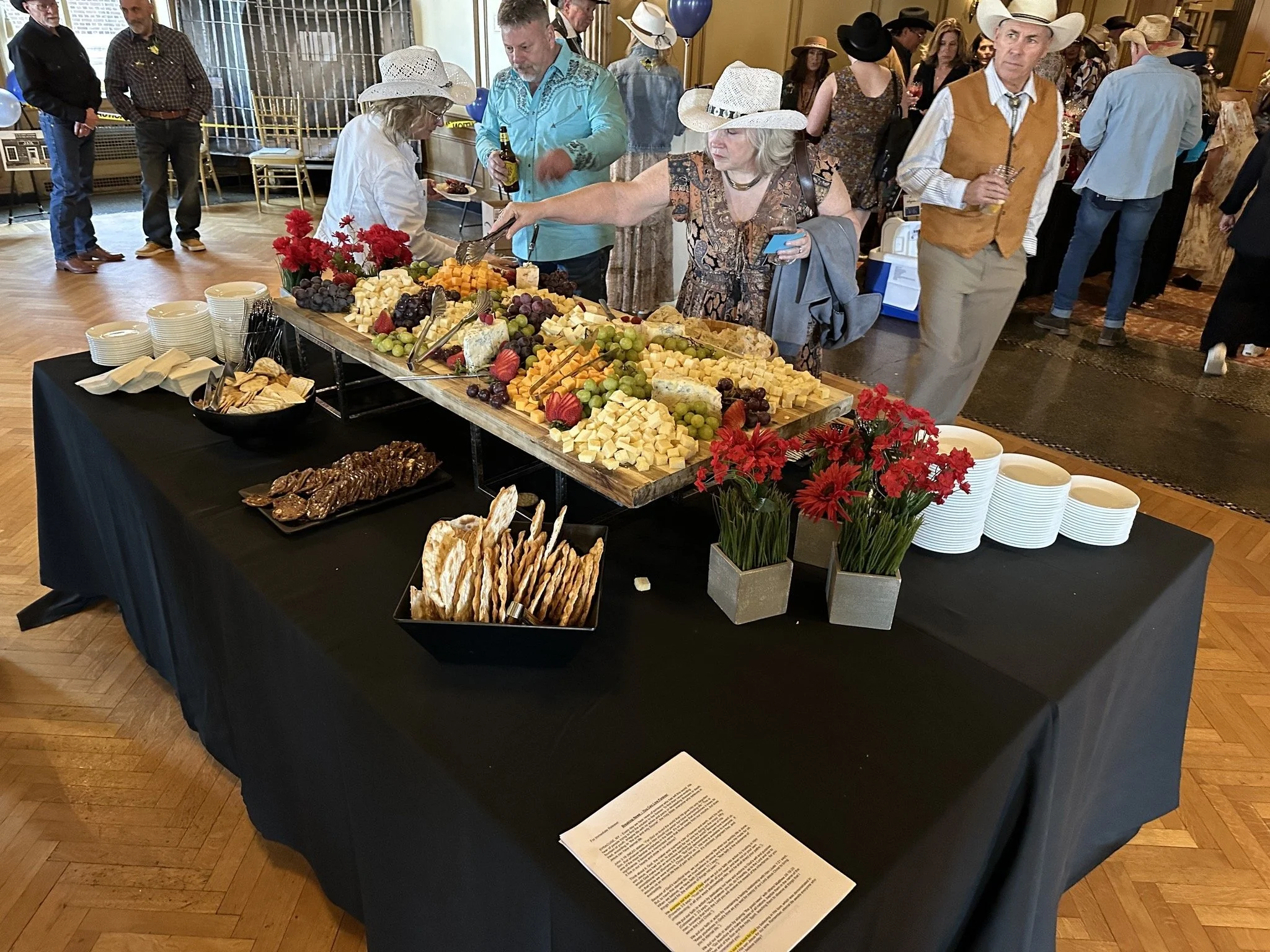 A buffet table with an assortment of cheeses, grapes, and strawberries, decorated with red flowers, served at a social event with people wearing cowboy hats and casual attire.
