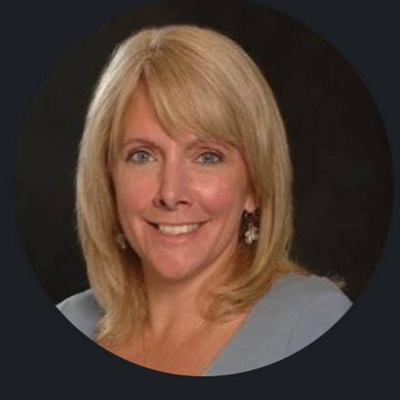 Headshot of a smiling woman with blonde hair and blue eyes, wearing earrings and a light-colored top, against a dark background.