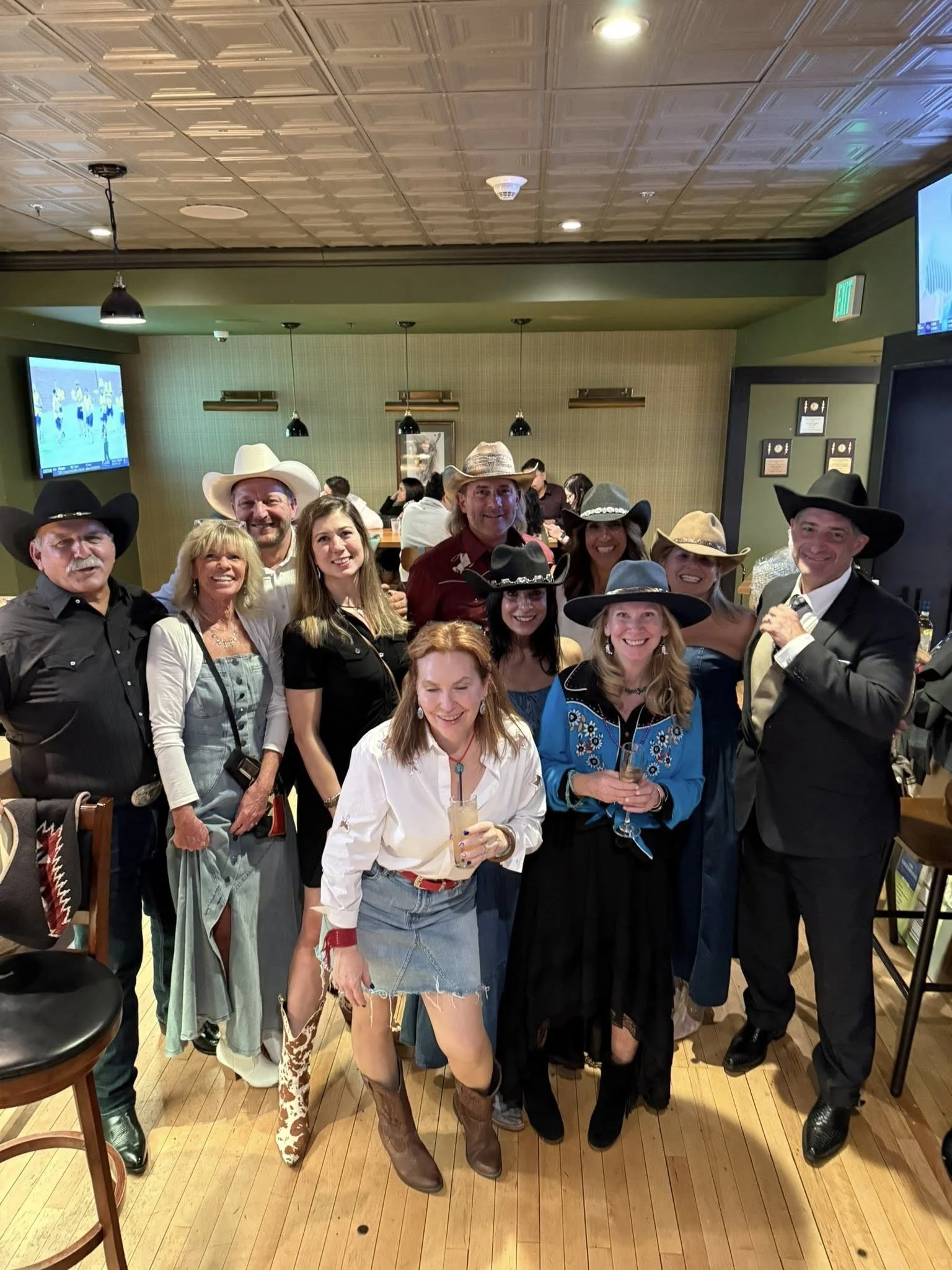 Group of people in cowboy hats at a social gathering, some holding drinks, inside a restaurant or bar.