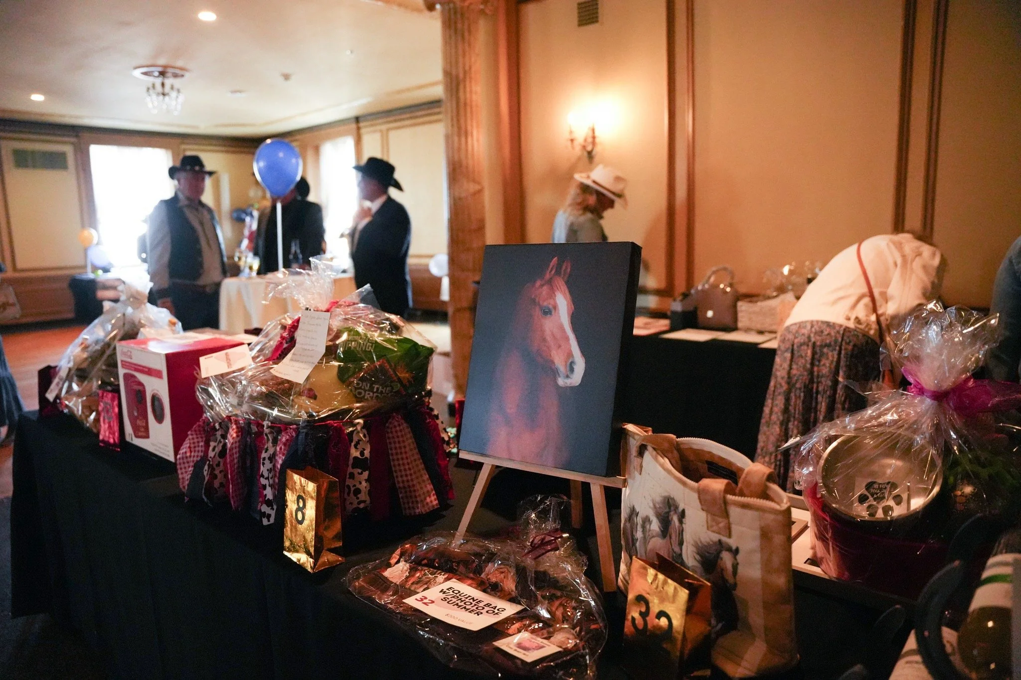 Auction table with wrapped gifts, a painting of a horse, and people in cowboy hats in a warmly lit room.