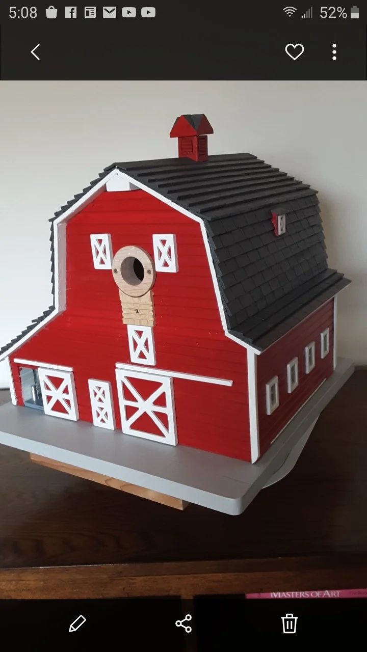 A detailed red and white miniature model of a barn with a black roof, small windows, a circular feature on the front, and a small red chimney on top, placed on a gray base.