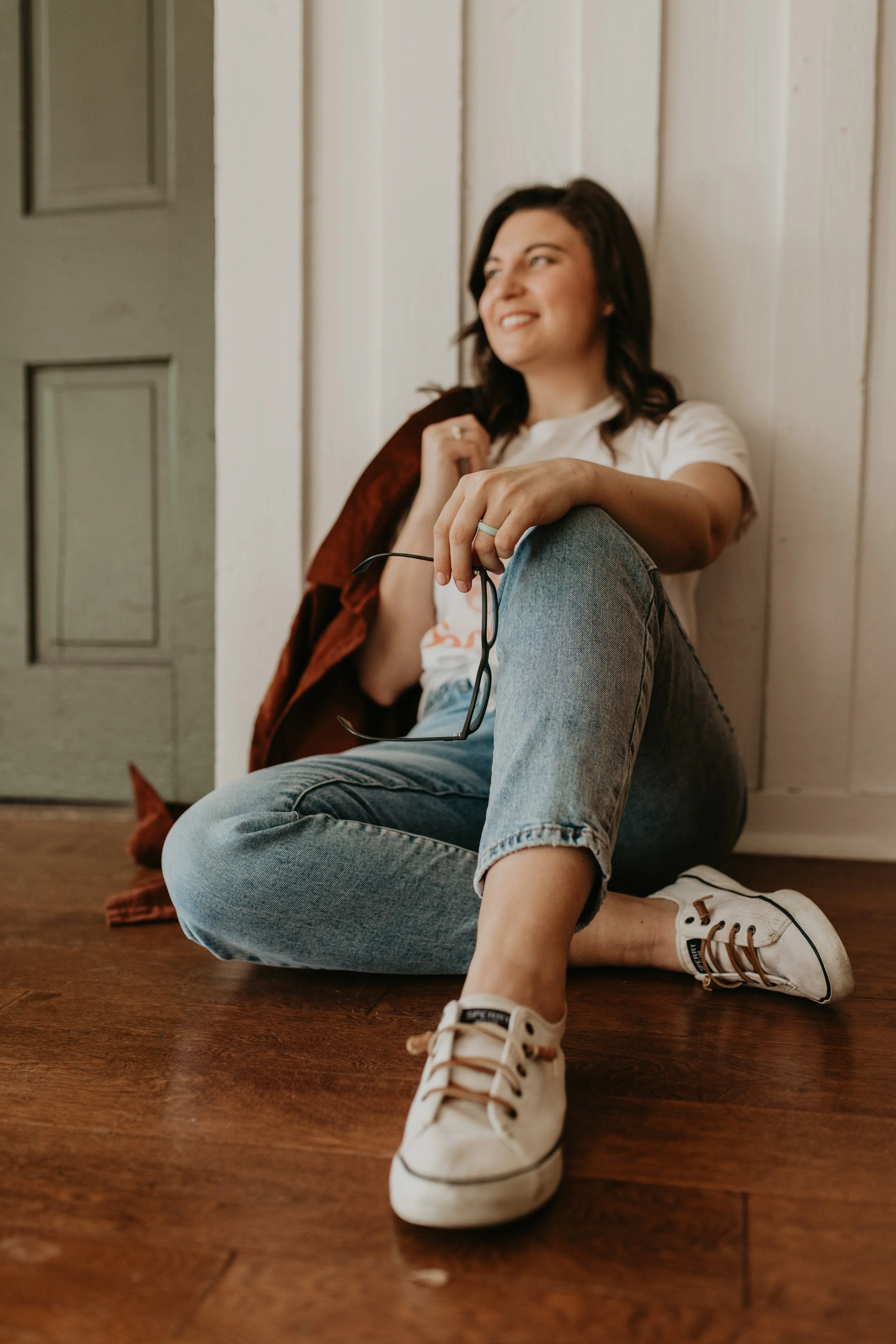 Female eating disorder therapist with brown hair, sitting on the floor with her leg propped up, holding her glasses and looking to her right side.