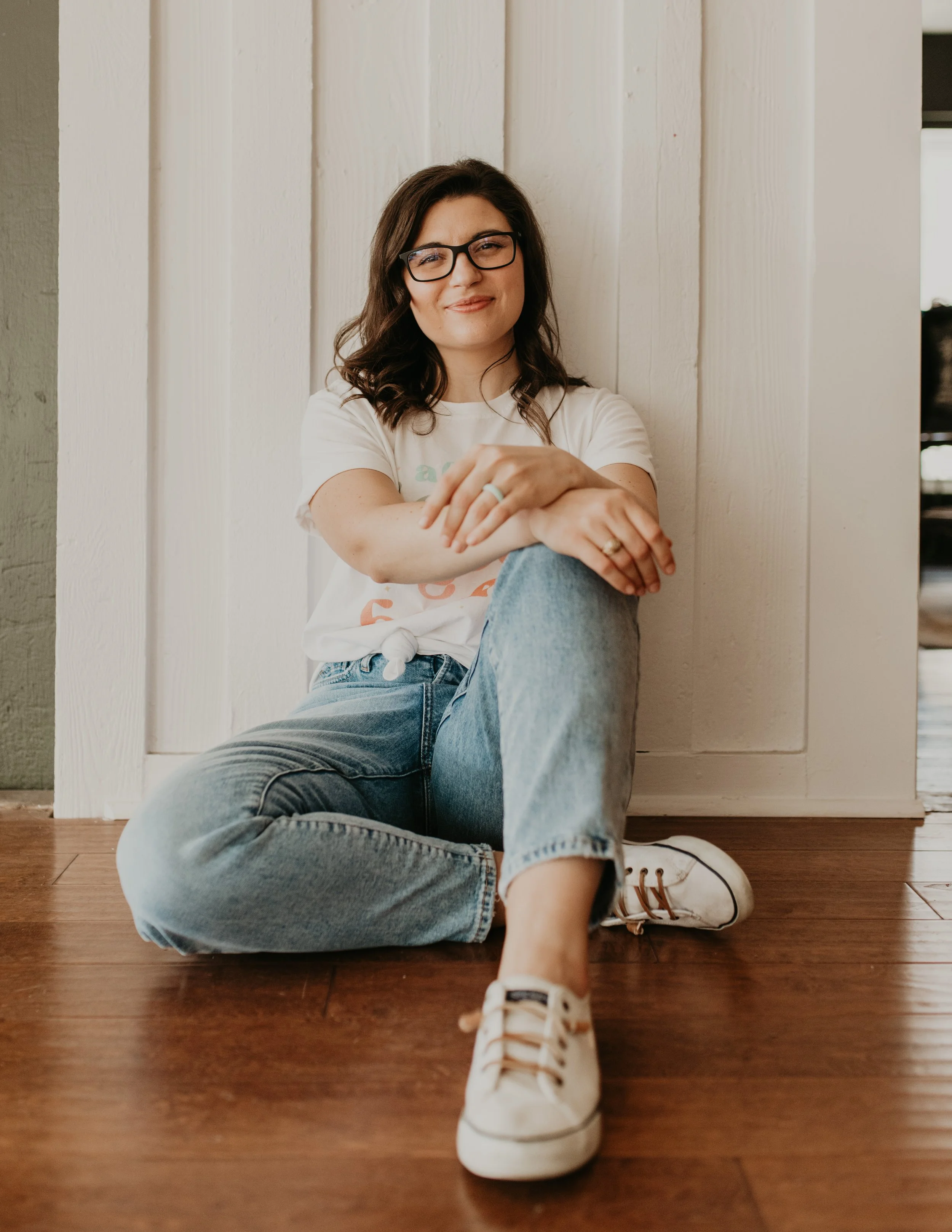 A female eating disorder and OCD therapist sitting on the floor with one legged propped up. She has brown hair and glasses. Her arms are crossed in a relaxed placement on her knee.
