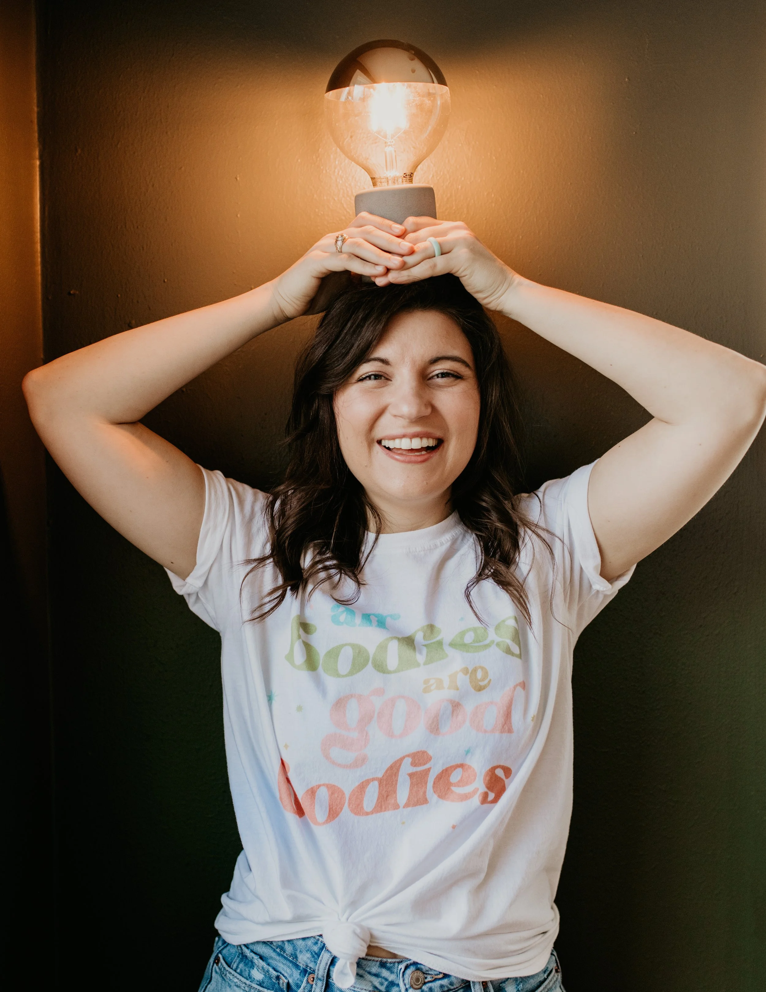 A woman with brown hair and a cheerful smile holds a lightbulb above her head, making it appear as if she has a lightbulb on her head, standing against a dark wall.