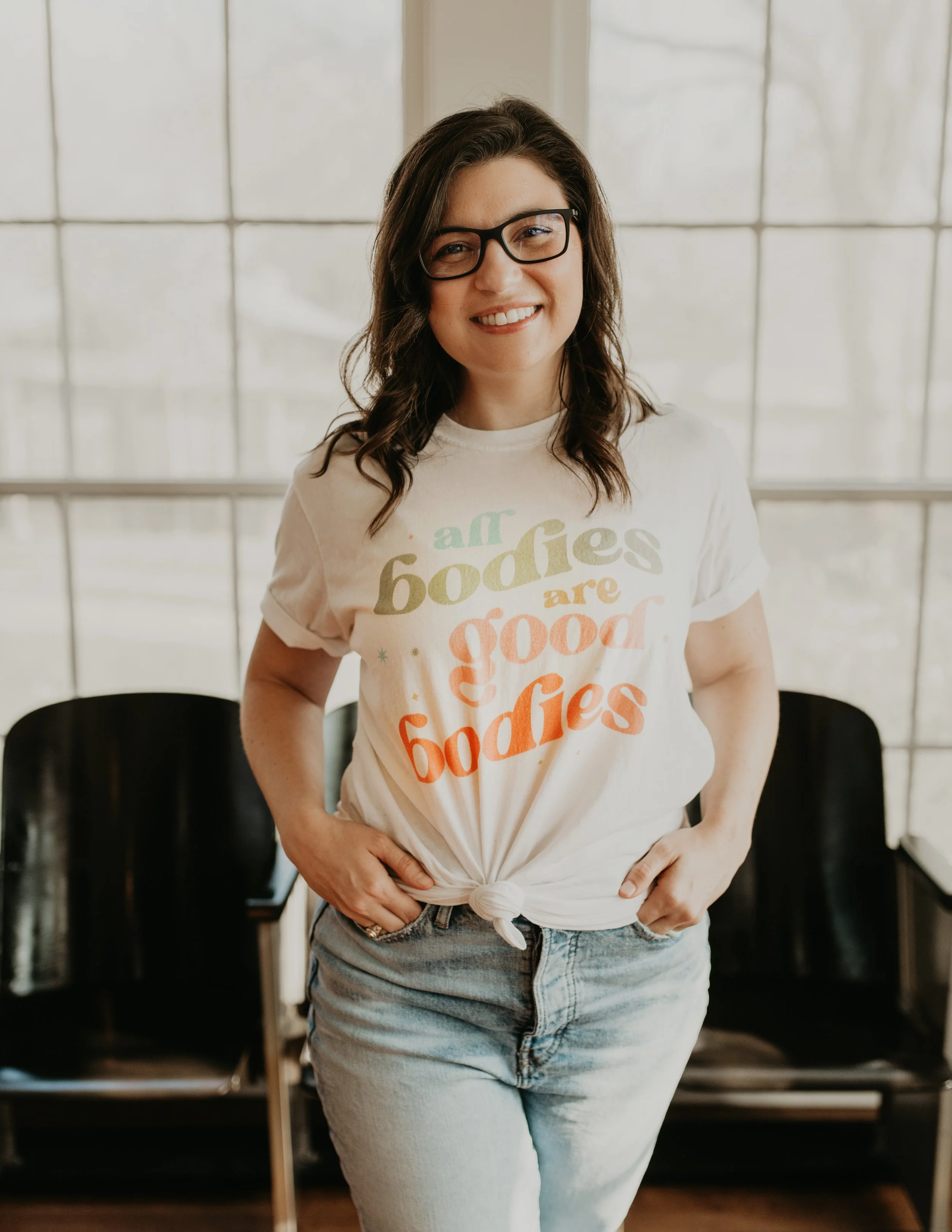 An OCD + Anxiety female therapist with brown hair and glasses standing with her hands in her jean pockets. Her shirt says "all bodies are good bodies".
