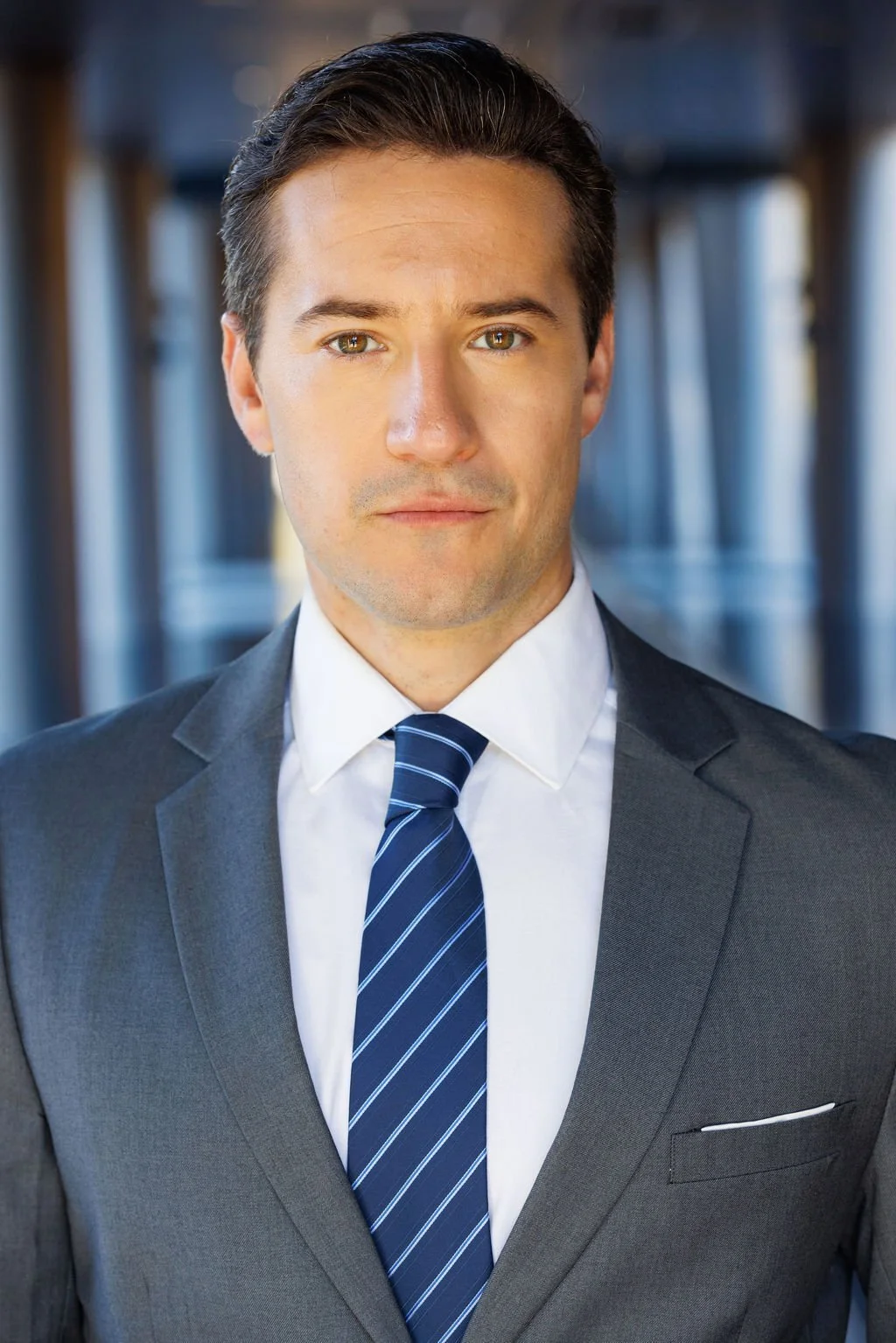 Portrait of a young businessman in a dark suit, white shirt, and blue striped tie, standing in an indoor corridor with large windows.