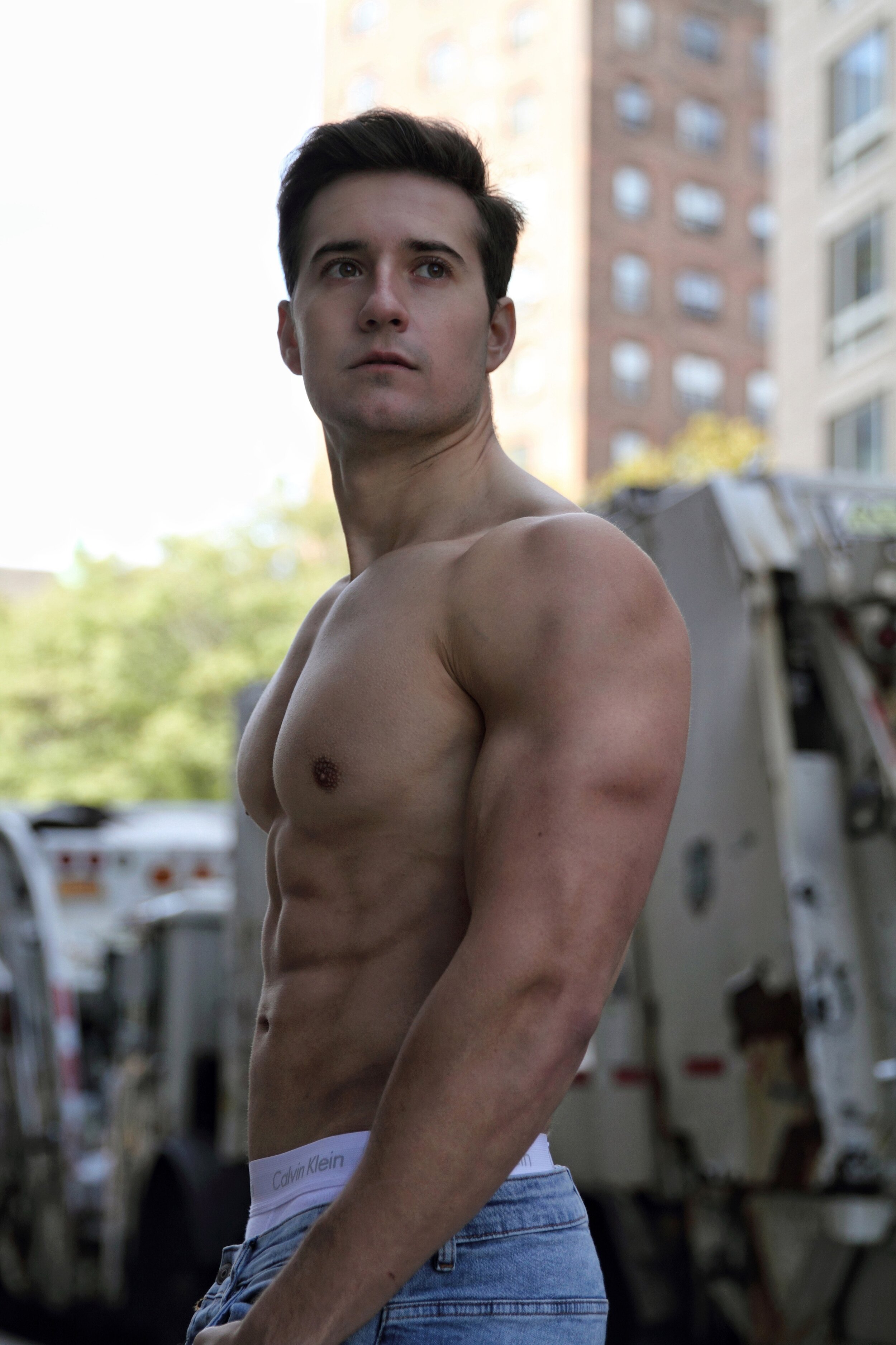 A shirtless young man with dark hair and light skin standing outdoors in an urban area with buildings and a dumpster in the background. He is wearing blue jeans and Calvin Klein underwear waistband visible.