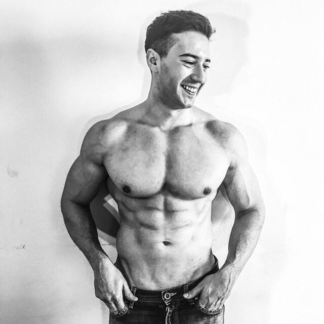 A shirtless man with a muscular physique standing against a plain wall, smiling and looking to his right.