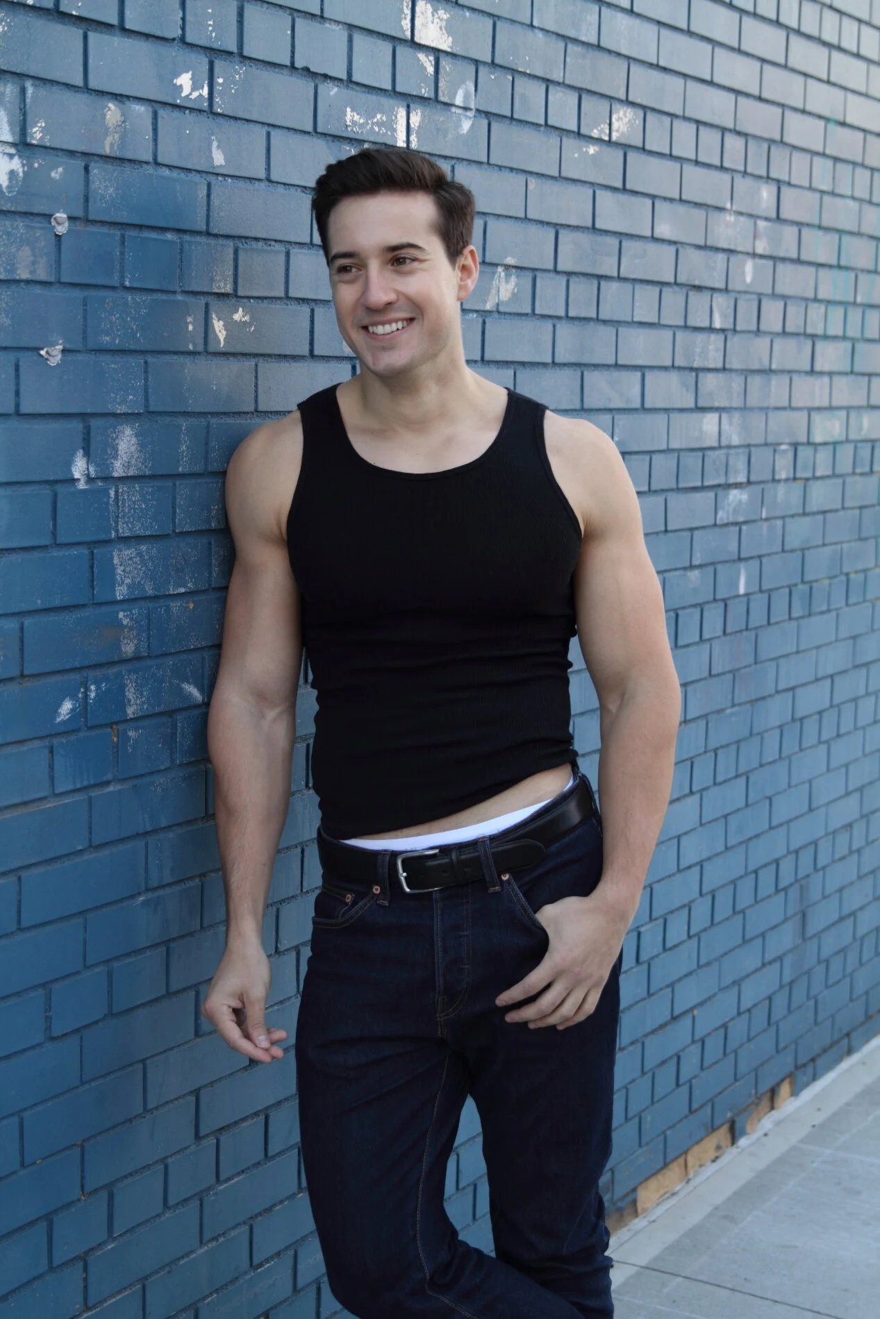 A young man with dark hair wearing a black tank top and dark jeans, standing against a blue brick wall with some peeled paint and smiling.