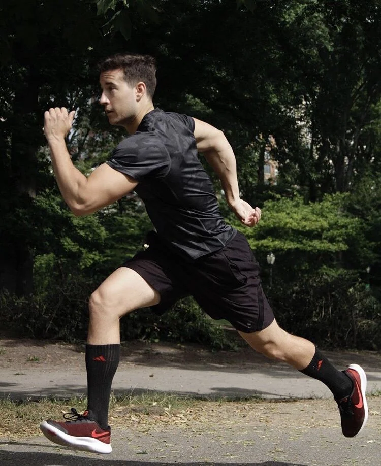 A man running outdoors in a park, wearing black athletic shorts, a black T-shirt, black socks, and red and black running shoes.