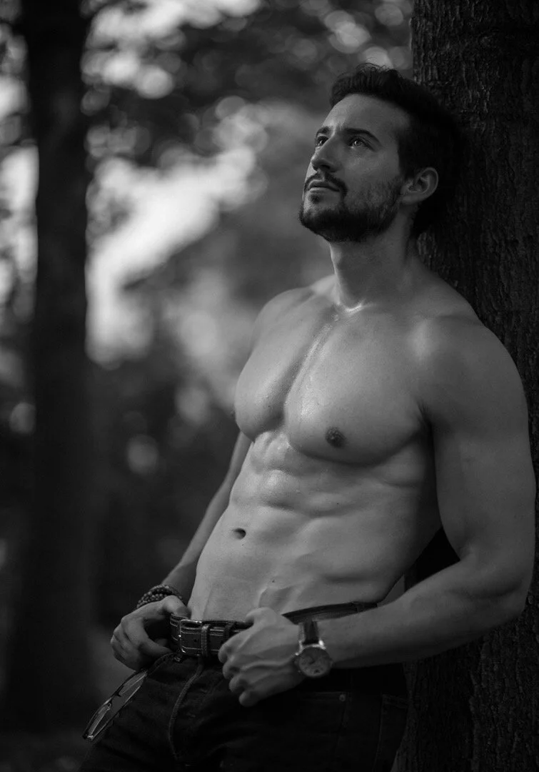 A shirtless man with a beard leaning against a tree in a forest, looking upward with a contemplative expression.