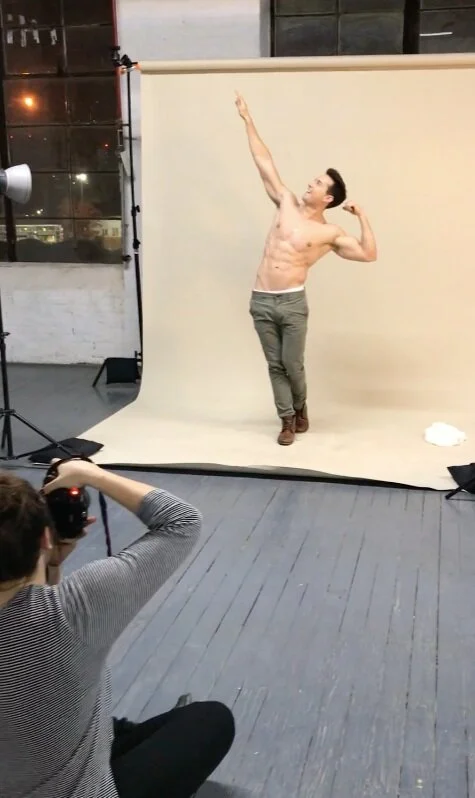 A shirtless man is posing confidently with one arm raised and the other flexed, standing on a photography studio backdrop while a photographer captures his picture.